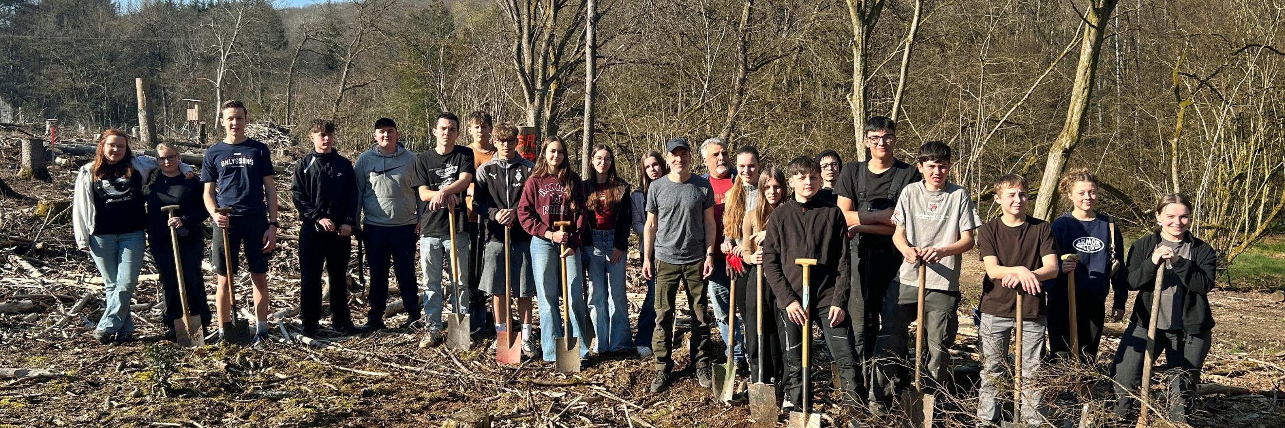 Eine große Gruppe Jugendlicher und Erwachsener steht auf einer offenen, kahlen Waldfläche zwischen dünnen, blattlosen Bäumen. Der Boden ist mit Ästen, Rindenschollen und Holzresten bedeckt. Viele der Teilnehmenden halten Spaten in den Händen und stehen in einer Reihe für ein Gruppenfoto. Im Hintergrund sind mehr Bäume, sanfte Hügel und ein blauer Himmel mit etwas Sonnenschein zu sehen.