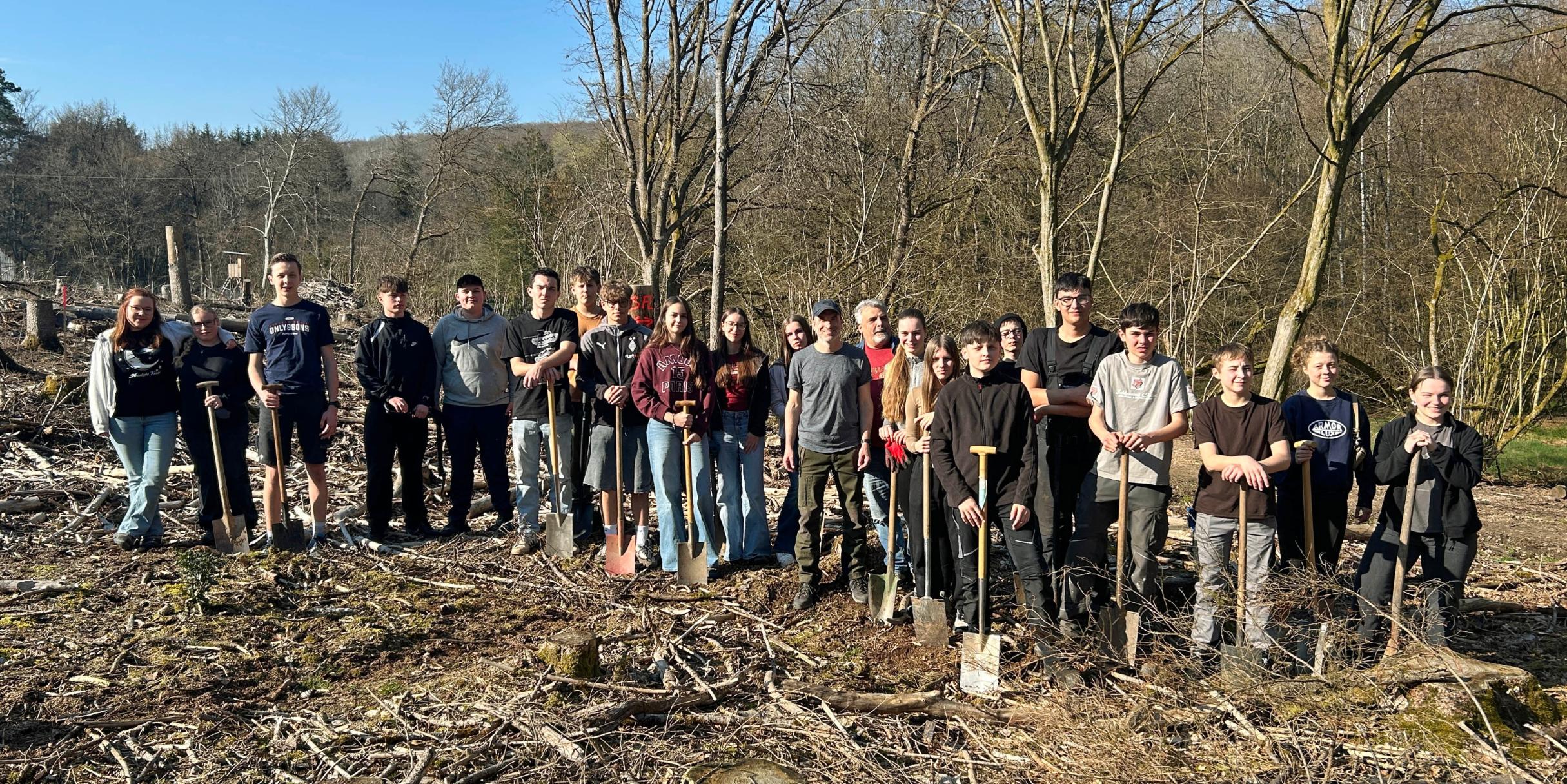 Eine große Gruppe Jugendlicher und Erwachsener steht auf einer offenen, kahlen Waldfläche zwischen dünnen, blattlosen Bäumen. Der Boden ist mit Ästen, Rindenschollen und Holzresten bedeckt. Viele der Teilnehmenden halten Spaten in den Händen und stehen in einer Reihe für ein Gruppenfoto. Im Hintergrund sind mehr Bäume, sanfte Hügel und ein blauer Himmel mit etwas Sonnenschein zu sehen.