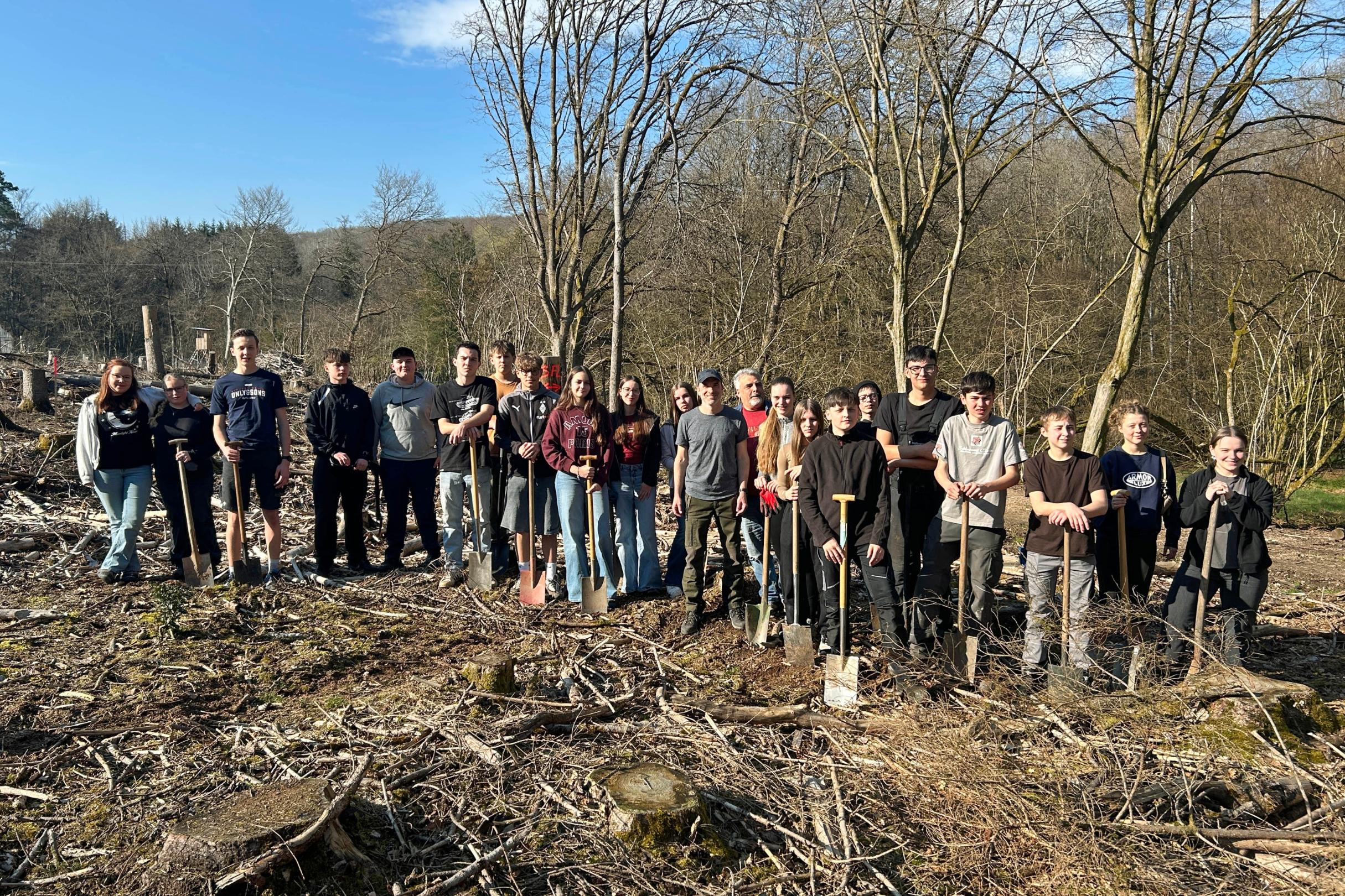 Eine große Gruppe Jugendlicher und Erwachsener steht auf einer offenen, kahlen Waldfläche zwischen dünnen, blattlosen Bäumen. Der Boden ist mit Ästen, Rindenschollen und Holzresten bedeckt. Viele der Teilnehmenden halten Spaten in den Händen und stehen in einer Reihe für ein Gruppenfoto. Im Hintergrund sind mehr Bäume, sanfte Hügel und ein blauer Himmel mit etwas Sonnenschein zu sehen.