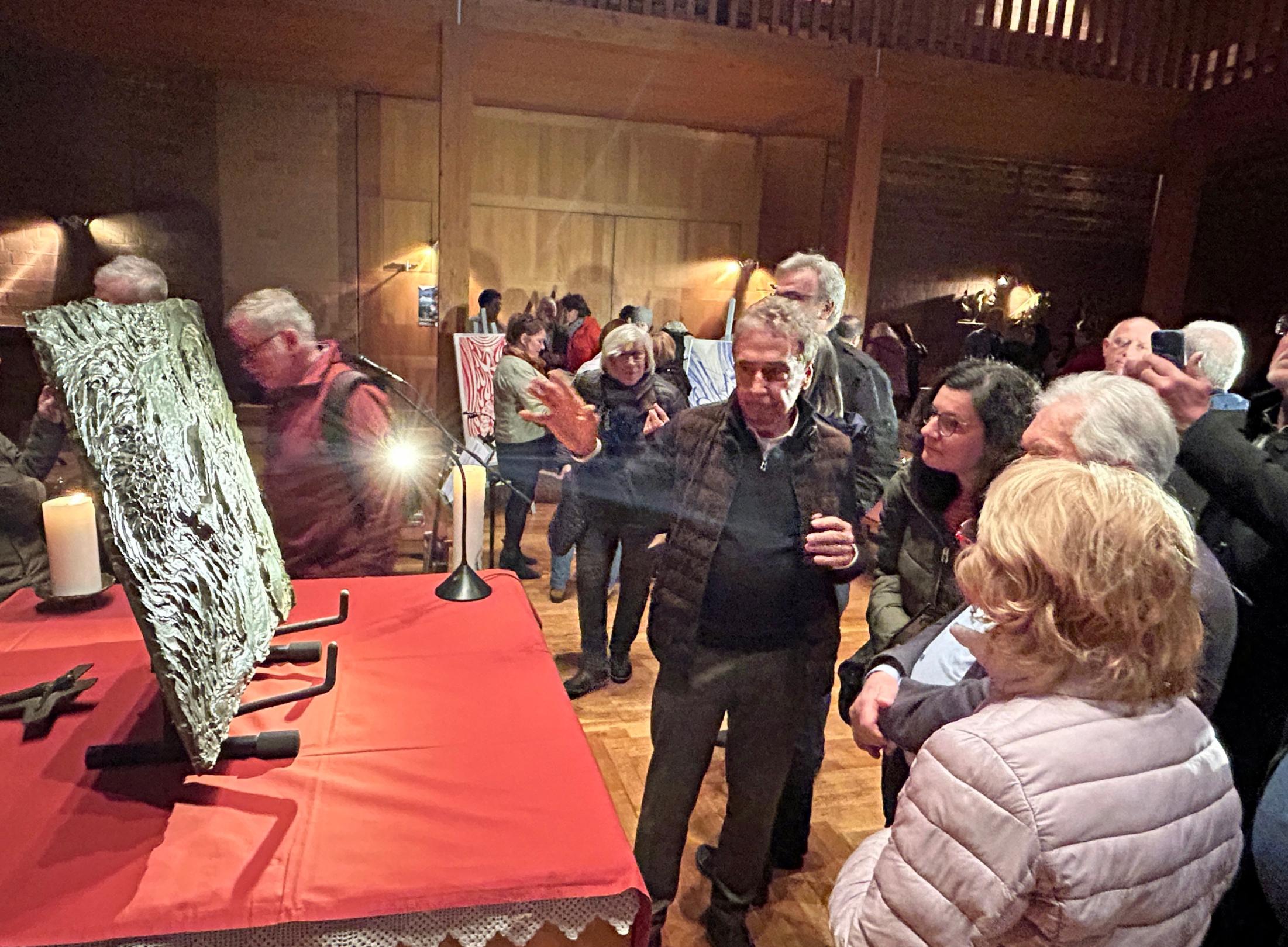 Im Innenraum einer Kirche betrachten zahlreiche Besucher ausgestellte Kunstwerke. Im Vordergrund steht ein großer, strukturierter Bildträger auf einem rot gedeckten Tisch, beleuchtet von einer kleinen Lampe. Daneben brennt eine Kerze. Viele Menschen stehen dicht beisammen und schauen die Kunstwerke aus der Nähe an oder unterhalten sich darüber. Im Hintergrund sind weitere Bilder auf Staffeleien zu sehen sowie eine Holzempore und die holzverkleidete Wand der Kirche. Die Szene ist warm beleuchtet und vermittelt den Eindruck einer gut besuchten Ausstellung.