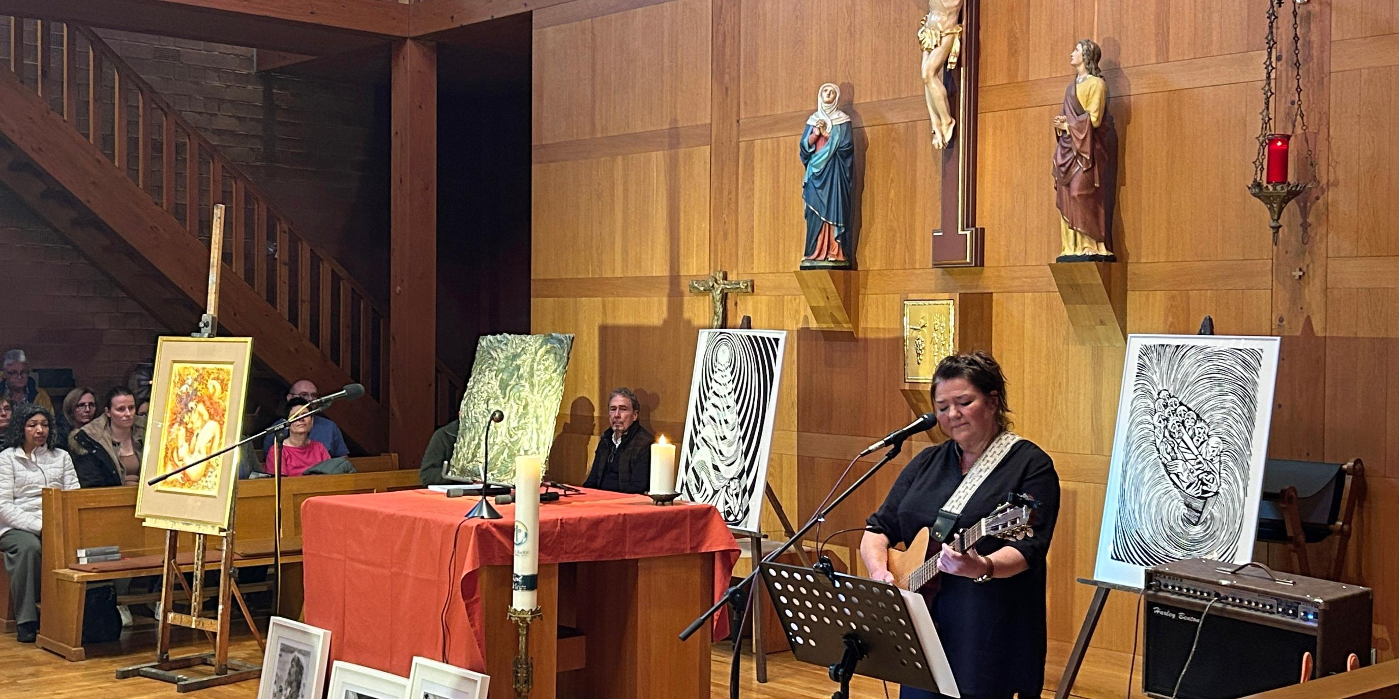 Innenraum einer Kirche mit holzverkleideten Wänden und einer erhöhten Empore im Hintergrund. Zentral an der Rückwand hängt ein großes Kruzifix, flankiert von zwei weiteren Heiligenfiguren. Vor dem Altarbereich stehen mehrere Staffeleien mit Kunstwerken, darunter farbige Gemälde und schwarz‑weiße Grafiken. Auf dem Boden vor dem Altar sind weitere gerahmte Bilder aufgereiht. Der Altar ist mit einem roten Tuch bedeckt, darauf stehen Kerzen und Mikrofone. Rechts im Vordergrund spielt eine Person Gitarre und singt in ein Mikrofon. Links und im Hintergrund sitzen Besucher in Kirchenbänken und verfolgen die Veranstaltung. Die Beleuchtung ist warm und lässt den Holzraum klar hervortreten.