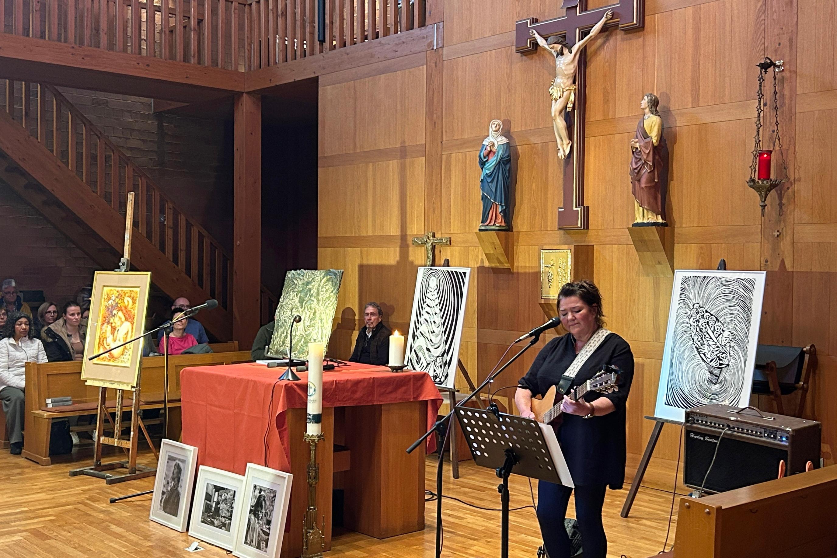 Innenraum einer Kirche mit holzverkleideten Wänden und einer erhöhten Empore im Hintergrund. Zentral an der Rückwand hängt ein großes Kruzifix, flankiert von zwei weiteren Heiligenfiguren. Vor dem Altarbereich stehen mehrere Staffeleien mit Kunstwerken, darunter farbige Gemälde und schwarz‑weiße Grafiken. Auf dem Boden vor dem Altar sind weitere gerahmte Bilder aufgereiht. Der Altar ist mit einem roten Tuch bedeckt, darauf stehen Kerzen und Mikrofone. Rechts im Vordergrund spielt eine Person Gitarre und singt in ein Mikrofon. Links und im Hintergrund sitzen Besucher in Kirchenbänken und verfolgen die Veranstaltung. Die Beleuchtung ist warm und lässt den Holzraum klar hervortreten.