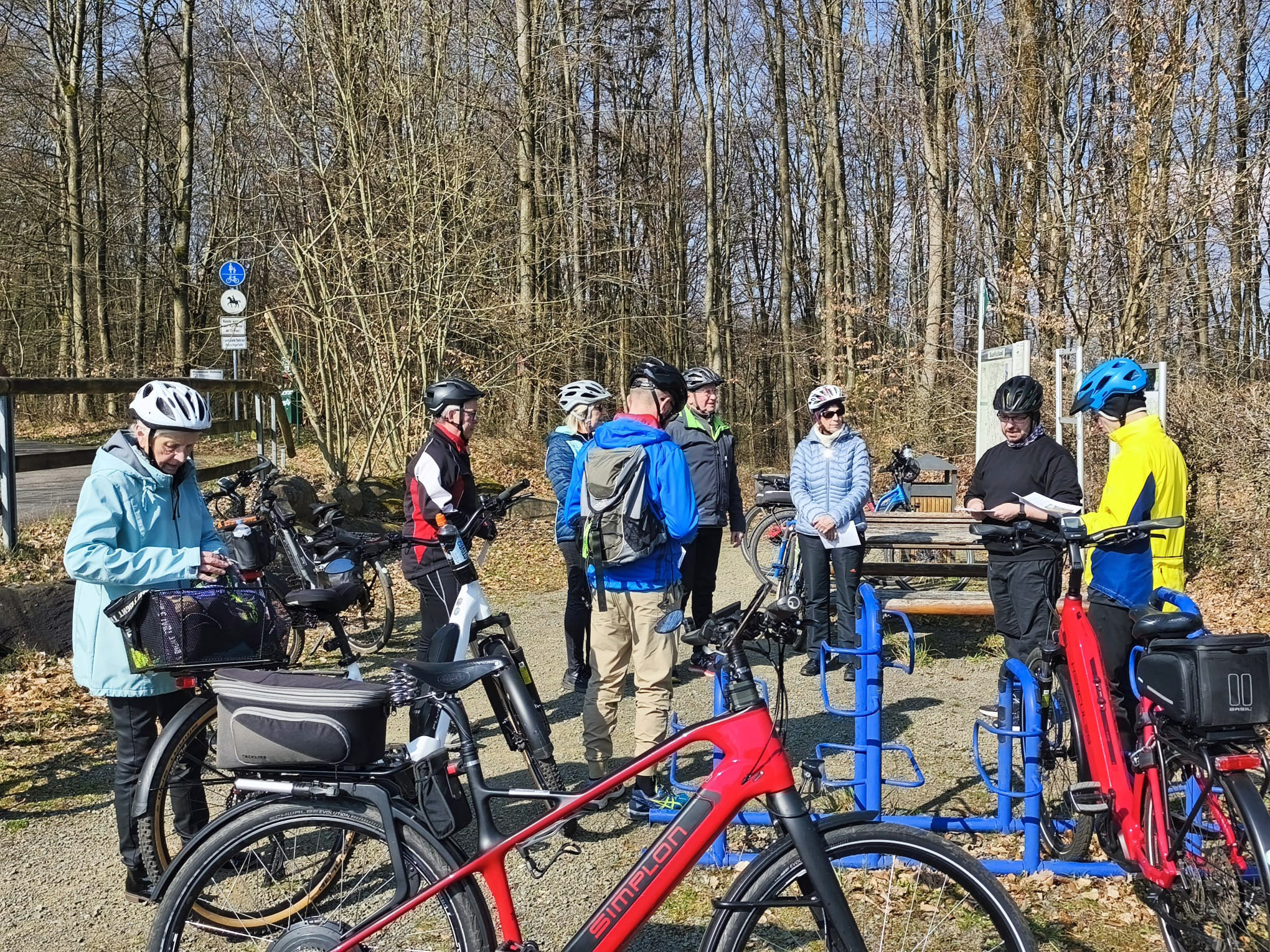 Eine Gruppe von mehreren Personen mit Fahrradhelmen steht an einem kleinen Rastplatz im Wald. Um sie herum stehen zahlreiche Fahrräder, darunter auch E‑Bikes, die teils an blauen Metallständern angelehnt sind. Die Personen tragen wetterfeste Kleidung und wirken damit für eine Fahrradtour ausgerüstet. Im Hintergrund stehen dicht gewachsene, laublose Bäume. Auf der linken Seite führt ein schmaler Weg vorbei, daneben befindet sich ein Verkehrszeichen für einen Radweg. Rechts im Hintergrund steht eine Holzbank. Die Szene ist sonnig und klar, mit deutlichen Schatten auf dem Boden. Geben Sie Ihr Feedback zu BizChat ab.