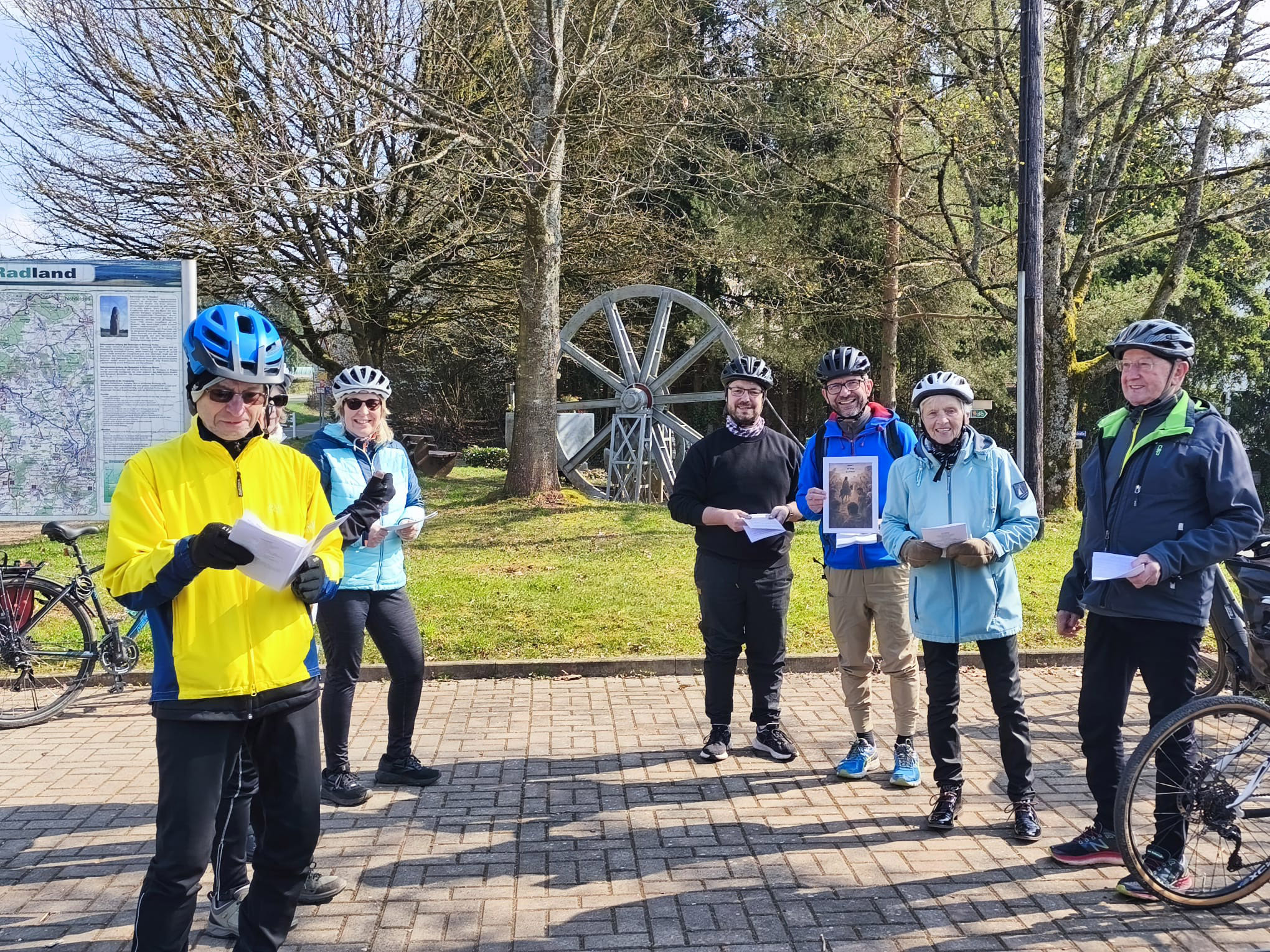 Eine Gruppe von sechs Personen steht mit Fahrrädern und Fahrradhelmen auf einem gepflasterten Platz im Freien. Einige halten Blätter oder Ausdrucke in den Händen. Im Hintergrund befinden sich Bäume, ein großes metallisches Mühlrad sowie eine Infotafel mit einer Karte. Das Wetter wirkt sonnig und klar, und mehrere Fahrräder lehnen am Rand des Platzes.