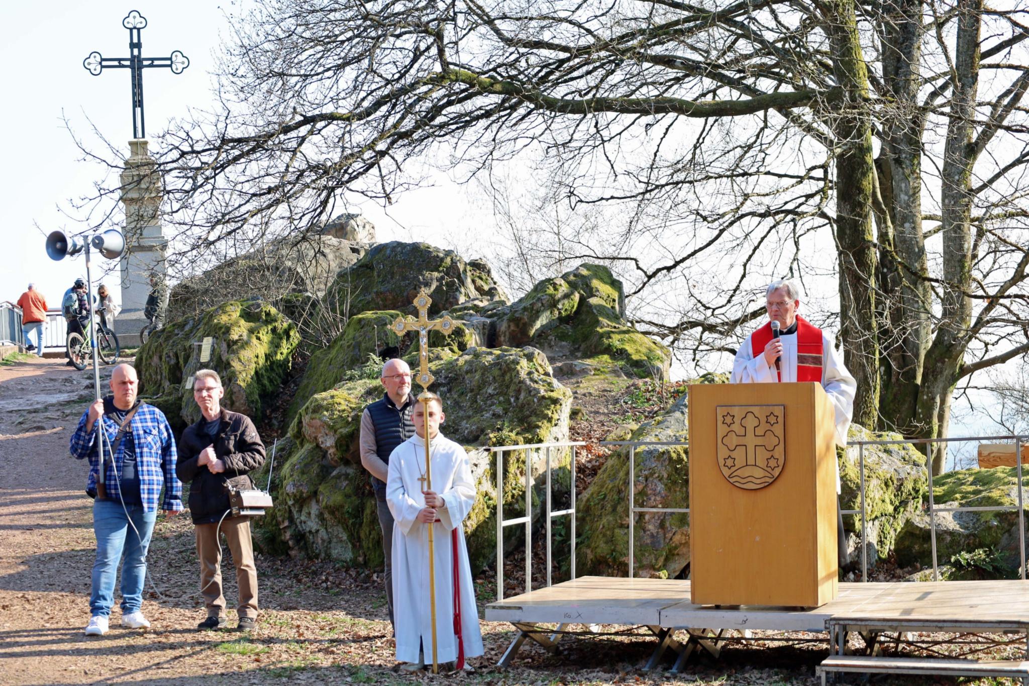 Auf einer kleinen erhöhten Holzplattform steht eine Person hinter einem Rednerpult, das mit einem Wappen versehen ist. Die Person trägt liturgische Gewänder und spricht zu einer Gruppe von Menschen, die außerhalb des Bildausschnitts stehen. Rechts und links der Plattform liegen große moosbewachsene Felsblöcke. Vor der Bühne stehen zwei Personen in weißen liturgischen Gewändern, eine davon hält ein Vortragekreuz. Weitere Personen stehen etwas seitlich daneben. Links im Hintergrund führt ein Weg zum Gipfelkreuz, das auf einem Felsen installiert ist und über eine kurze Treppe erreichbar ist. Entlang des Weges stehen mehrere Zuschauende sowie ein Lautsprecher. Die Szene spielt im Freien bei klarem Wetter, umgeben von kahlen Bäumen und natürlicher Felslandschaft.Geben Sie Ihr Feedback zu BizChat ab.
