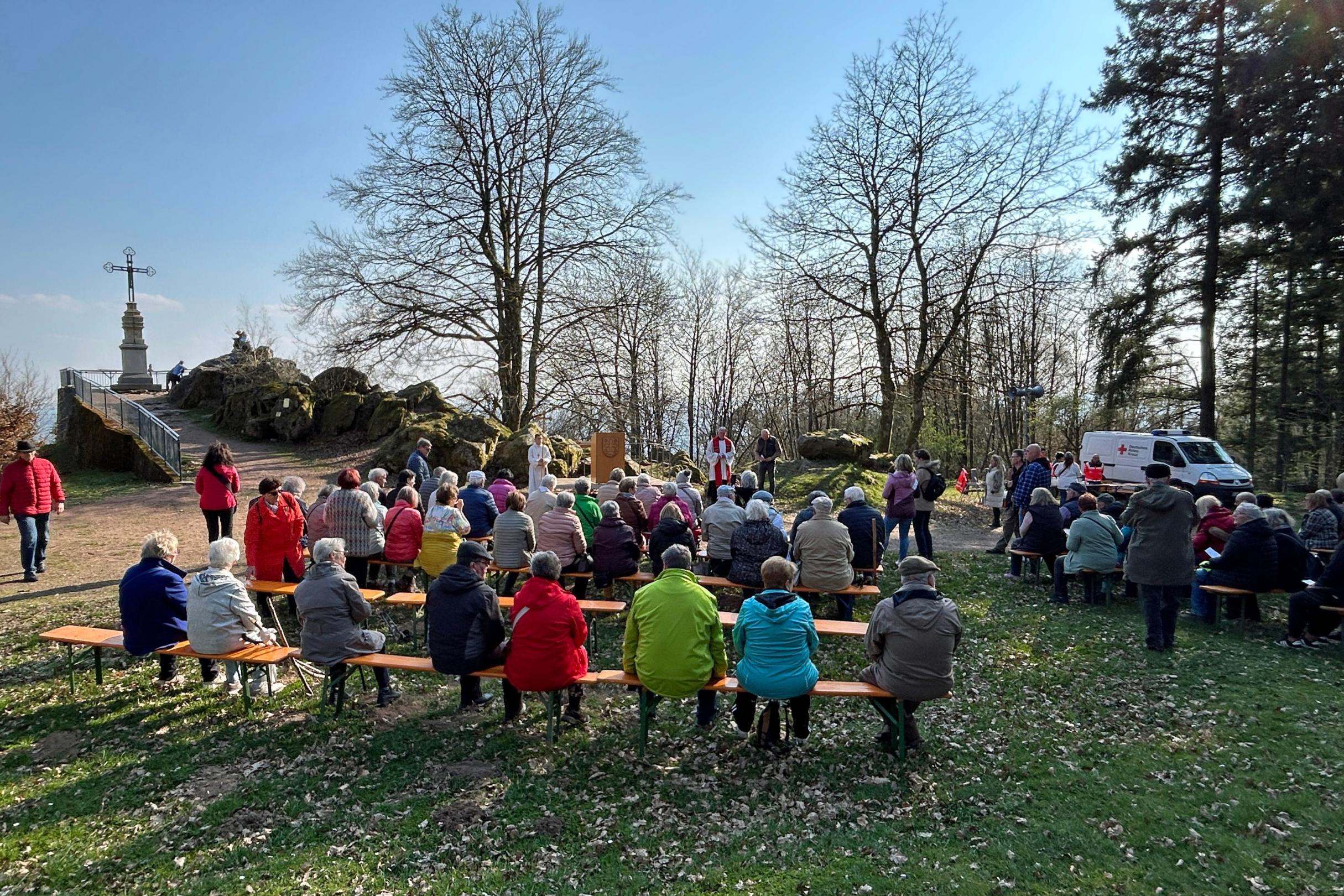 Auf einer Wiese am Rand eines bewaldeten Hügels sitzen zahlreiche Menschen auf einfachen Holzbänken, die in mehreren Reihen aufgestellt sind. Sie blicken nach vorn zu einer kleinen Freifläche, auf der ein Wortgottesdienst stattfindet. Dort stehen mehrere Personen in liturgischen Gewändern neben einem aufgestellten Kreuz. Rechts im Hintergrund steht ein Fahrzeug mit dem Logo eines Hilfsdienstes. Links ragt ein großes Gipfelkreuz auf einem Felsenplateau in die Höhe, das über eine kurze Treppe erreichbar ist. Um die Versammelten herum stehen noch kahle Bäume, und der Boden ist mit trockenem Laub bedeckt. Die Szene findet bei hellem Sonnenlicht unter blauem Himmel statt.