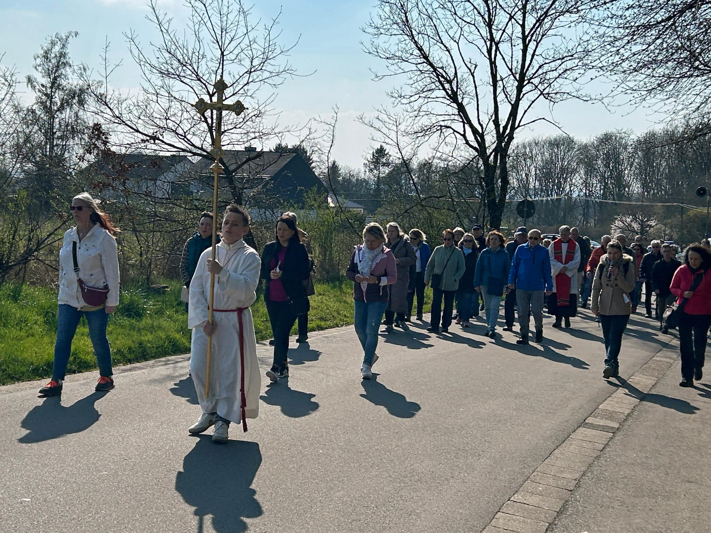 Eine größere Gruppe von Menschen geht bei sonnigem Wetter auf einer asphaltierten Straße zwischen Wiesen und Bäumen. An der Spitze der Prozession trägt eine Person ein großes Vortragekreuz. Hinter dieser Person folgt eine dicht geschlossene Gruppe von Teilnehmenden, einige in Jacken, andere mit Taschen oder Rucksäcken. Rechts und links des Weges stehen noch kahle Bäume und frühe Frühlingssträucher. Im Hintergrund sind einige Häuser und ein freies Feld zu sehen. Die Szene wirkt wie eine gemeinschaftliche religiöse Prozession oder ein gemeinsamer Weg durch die ländliche Umgebung.