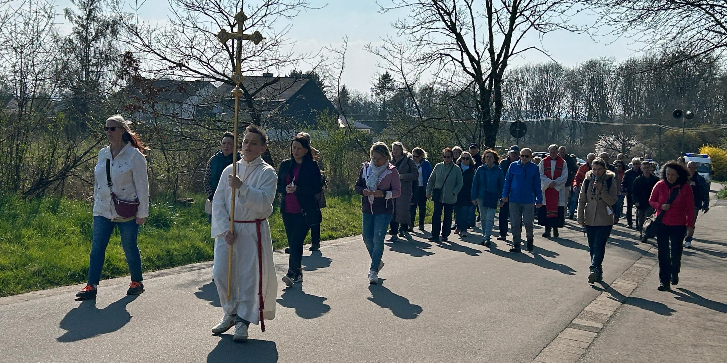 Eine größere Gruppe von Menschen geht bei sonnigem Wetter auf einer asphaltierten Straße zwischen Wiesen und Bäumen. An der Spitze der Prozession trägt eine Person ein großes Vortragekreuz. Hinter dieser Person folgt eine dicht geschlossene Gruppe von Teilnehmenden, einige in Jacken, andere mit Taschen oder Rucksäcken. Rechts und links des Weges stehen noch kahle Bäume und frühe Frühlingssträucher. Im Hintergrund sind einige Häuser und ein freies Feld zu sehen. Die Szene wirkt wie eine gemeinschaftliche religiöse Prozession oder ein gemeinsamer Weg durch die ländliche Umgebung.