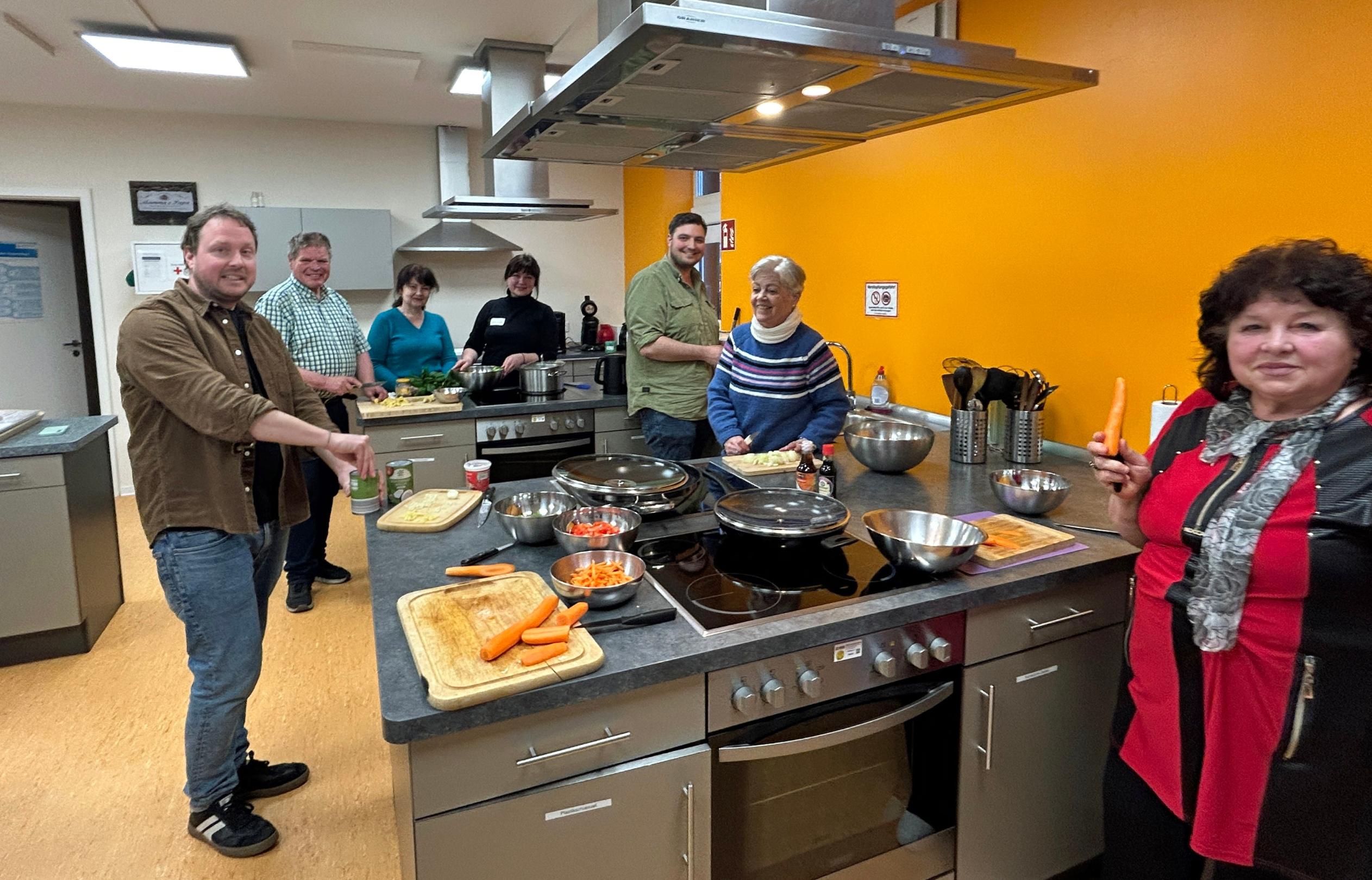 Eine Gruppe von sieben Menschen steht in einer Lehrküche und bereitet gemeinsam ein Essen zu. Mehrere Personen schneiden Gemüse, darunter Karotten, und lächeln in Richtung Kamera. Auf der zentralen Arbeitsfläche liegen Schneidebretter, Messer, Schüsseln und Pfannen mit bereits geschnittenem Gemüse. Die Küche ist modern ausgestattet, mit Edelstahlflächen, Kochfeldern und einer leuchtend orangefarbenen Wand im Hintergrund. Die Stimmung wirkt freundlich und gemeinschaftlich.