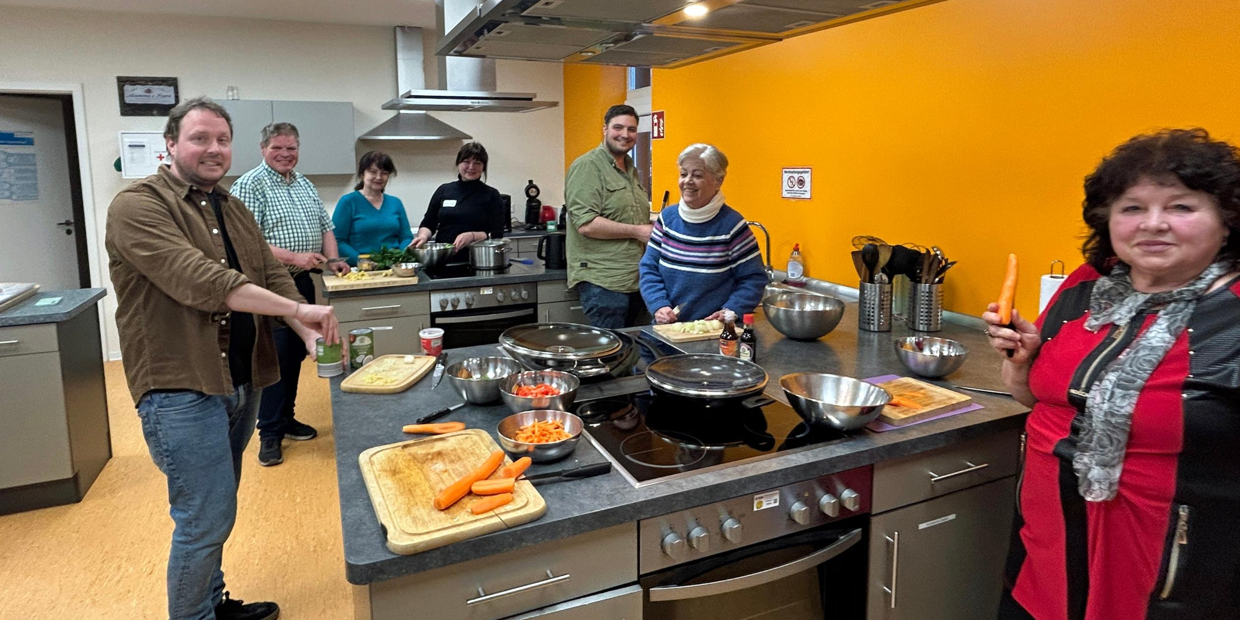 Eine Gruppe von sieben Menschen steht in einer Lehrküche und bereitet gemeinsam ein Essen zu. Mehrere Personen schneiden Gemüse, darunter Karotten, und lächeln in Richtung Kamera. Auf der zentralen Arbeitsfläche liegen Schneidebretter, Messer, Schüsseln und Pfannen mit bereits geschnittenem Gemüse. Die Küche ist modern ausgestattet, mit Edelstahlflächen, Kochfeldern und einer leuchtend orangefarbenen Wand im Hintergrund. Die Stimmung wirkt freundlich und gemeinschaftlich.