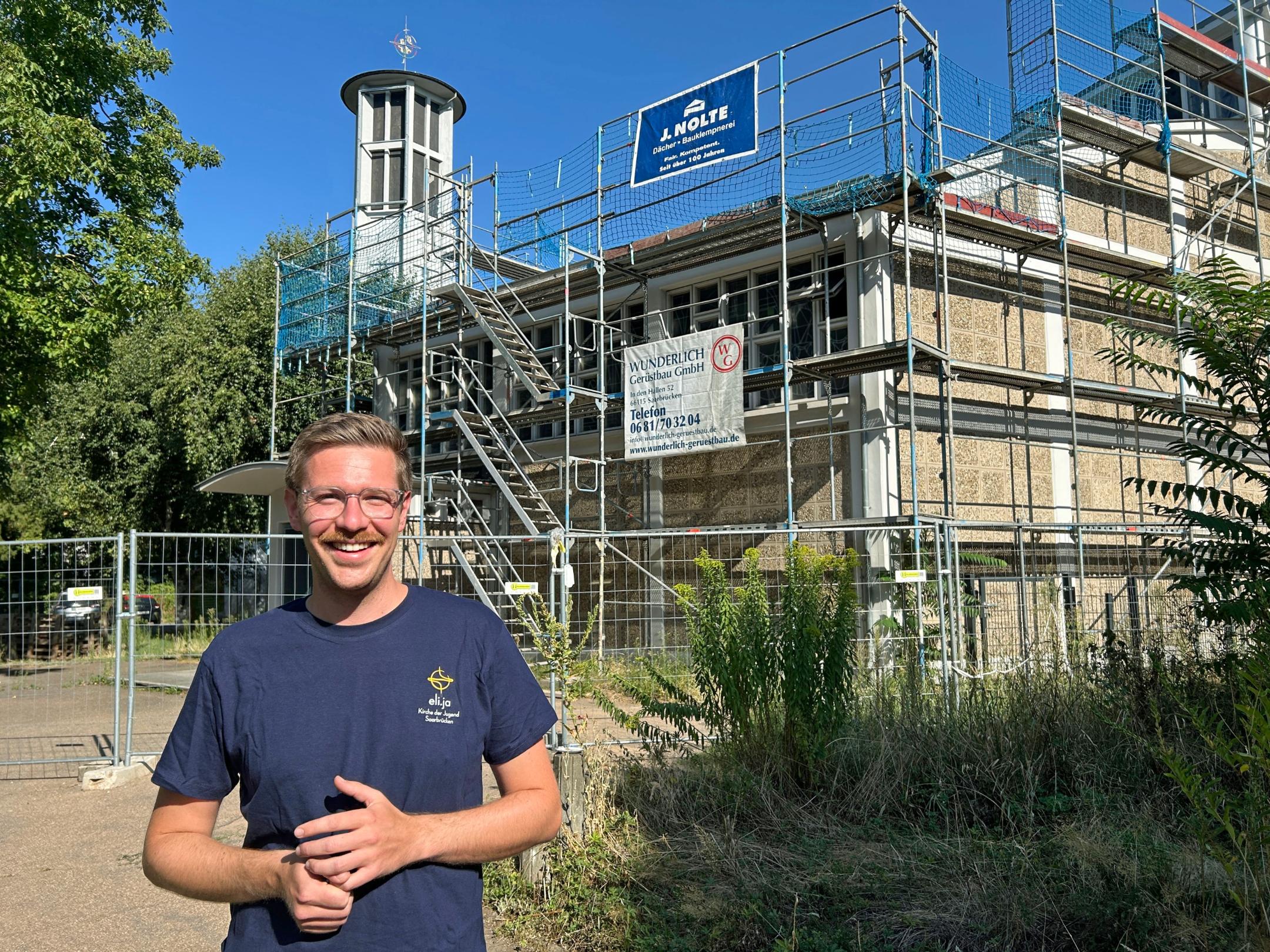 Ein Mann steht im Vordergrund auf einem Weg vor einem kirchlichen Gebäude. Das Gebäude ist vollständig von einem Gerüst mit Treppenaufgang umgeben und wird offenbar saniert. Hinter dem Gerüst ragt ein moderner Kirchturm mit rundem Dach und Kreuz in den blauen Himmel. Vor dem Gebäude stehen Bauzäune, am Gerüst hängen Werbebanner von Handwerksbetrieben. Links und rechts rahmen Bäume und Sträucher die Szene ein. Die Aufnahme entstand bei sonnigem Wetter mit klarem Himmel.