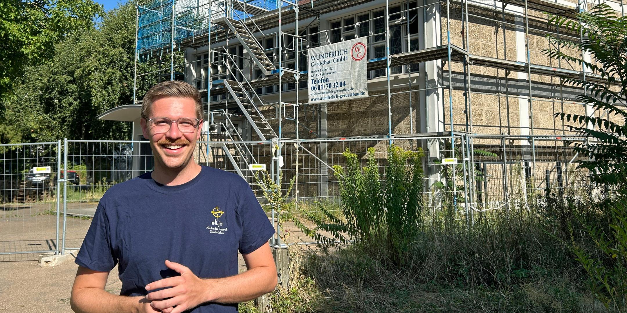 Patric Schützeichel (33) ist seit August der neue Saarbrücker Jugendpfarrer. Ein Mann steht im Vordergrund auf einem Weg vor einem kirchlichen Gebäude. Das Gebäude ist vollständig von einem Gerüst mit Treppenaufgang umgeben und wird offenbar saniert. Hinter dem Gerüst ragt ein moderner Kirchturm mit rundem Dach und Kreuz in den blauen Himmel. Vor dem Gebäude stehen Bauzäune, am Gerüst hängen Werbebanner von Handwerksbetrieben. Links und rechts rahmen Bäume und Sträucher die Szene ein. Die Aufnahme entstand bei sonnigem Wetter mit klarem Himmel.