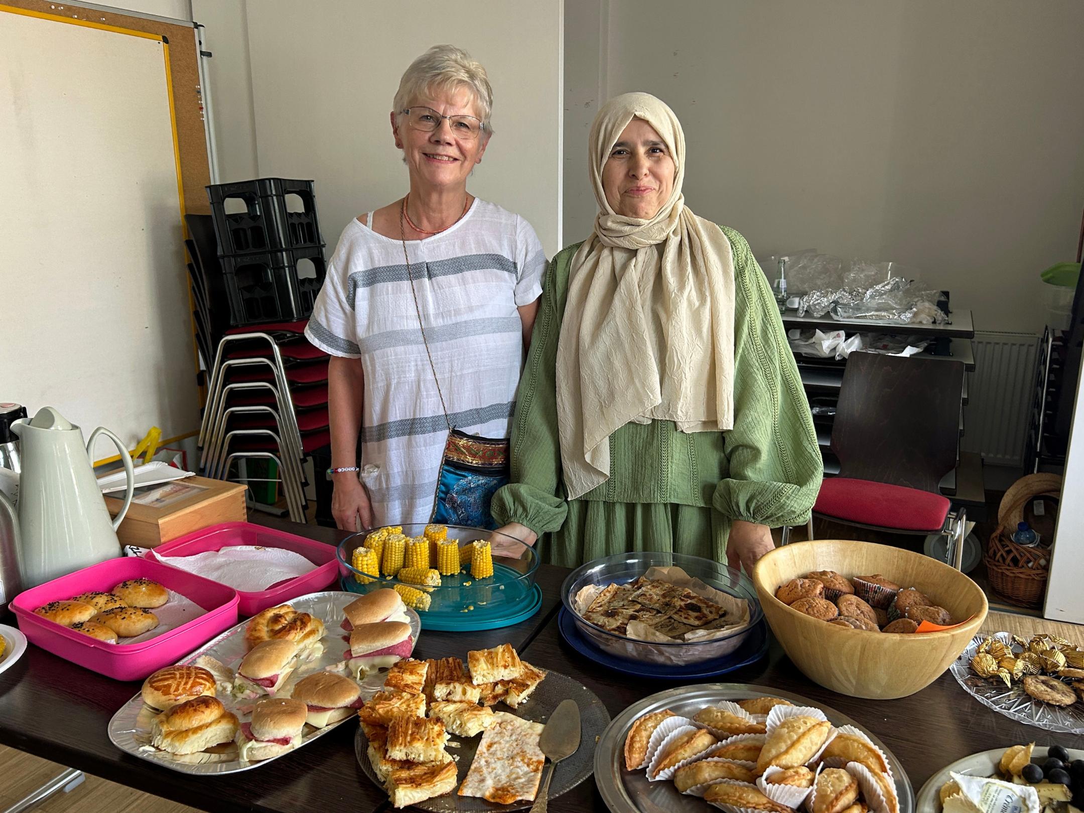 Zwei Frauen, eine Christin und eine Muslima mit Kopftuch, stehen hinter einem Buffet beim Friedensmahl des Interreligiösen Dialogkreises Völklingen. Auf dem Tisch stehen verschiedene Speisen, darunter gefüllte Brötchen, Gebäck, Maiskolben, Fladenbrot, Muffins und weitere internationale Spezialitäten.