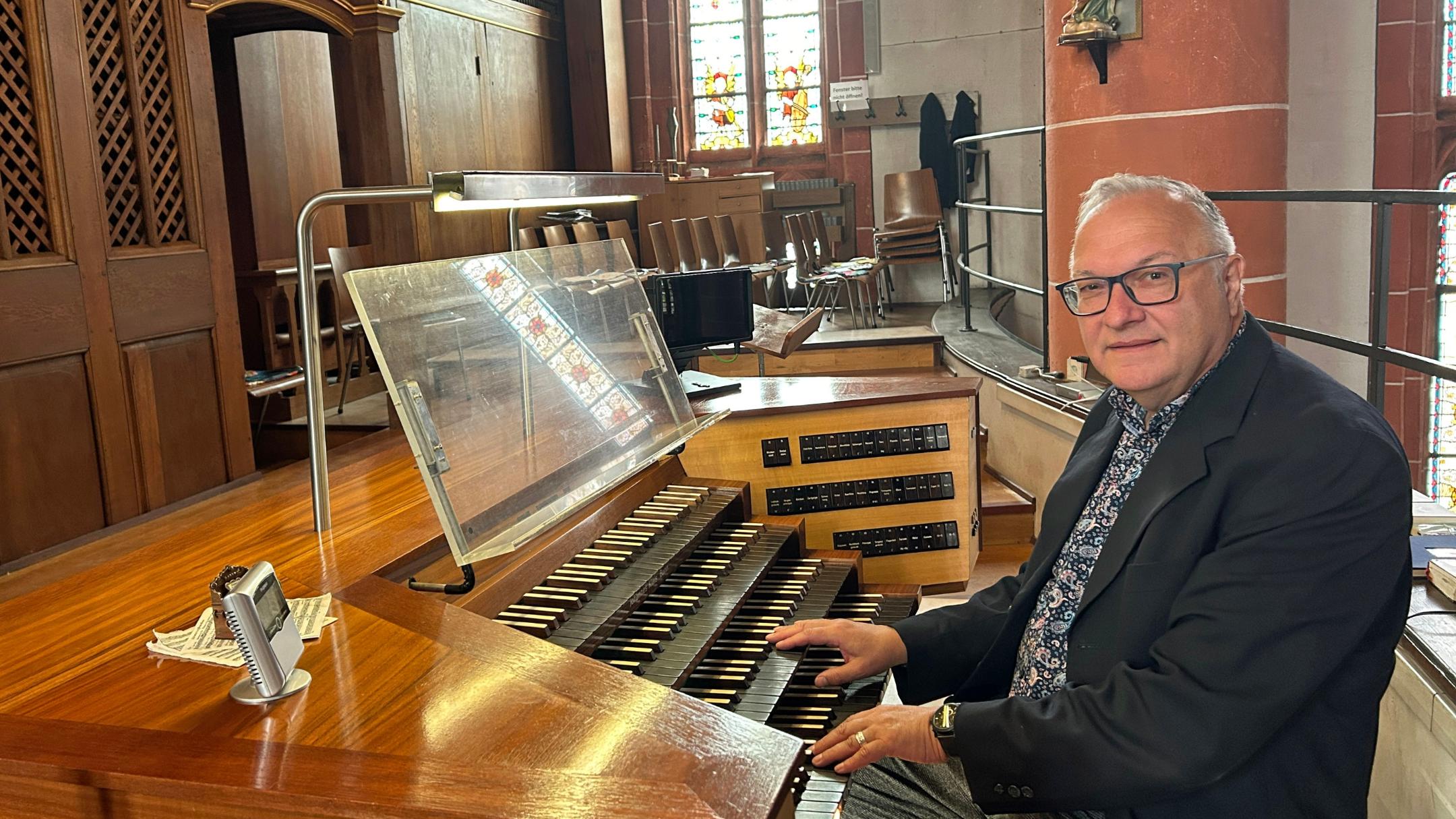 Ein Mann sitzt an der mehrmanualigen Orgel einer Kirche und hat die Hände auf den Tasten. Der hölzerne Spieltisch mit Notenpult und Registerschaltern steht auf einer Empore vor dem Orgelgehäuse mit sichtbaren Pfeifen. Im Hintergrund fallen Lichtstrahlen durch hohe Buntglasfenster, daneben sind Stuhlreihen und eine rötliche Steinsäule mit einer Heiligenfigur zu sehen. Die warme Holzoptik der Orgel prägt das Bild.