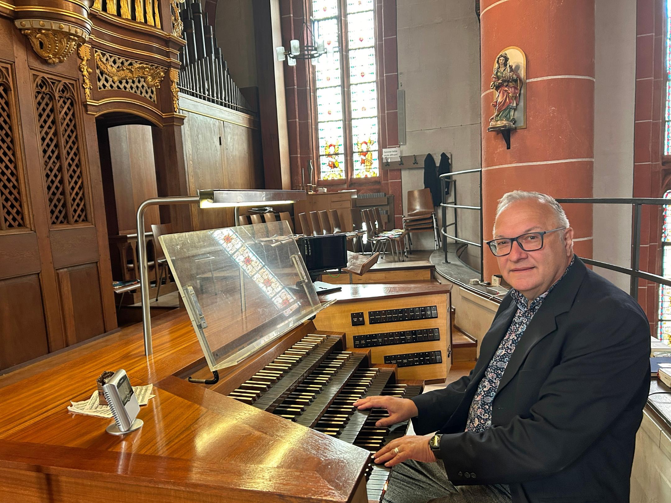 Ein Mann sitzt an der mehrmanualigen Orgel einer Kirche und hat die Hände auf den Tasten. Der hölzerne Spieltisch mit Notenpult und Registerschaltern steht auf einer Empore vor dem Orgelgehäuse mit sichtbaren Pfeifen. Im Hintergrund fallen Lichtstrahlen durch hohe Buntglasfenster, daneben sind Stuhlreihen und eine rötliche Steinsäule mit einer Heiligenfigur zu sehen. Die warme Holzoptik der Orgel prägt das Bild.