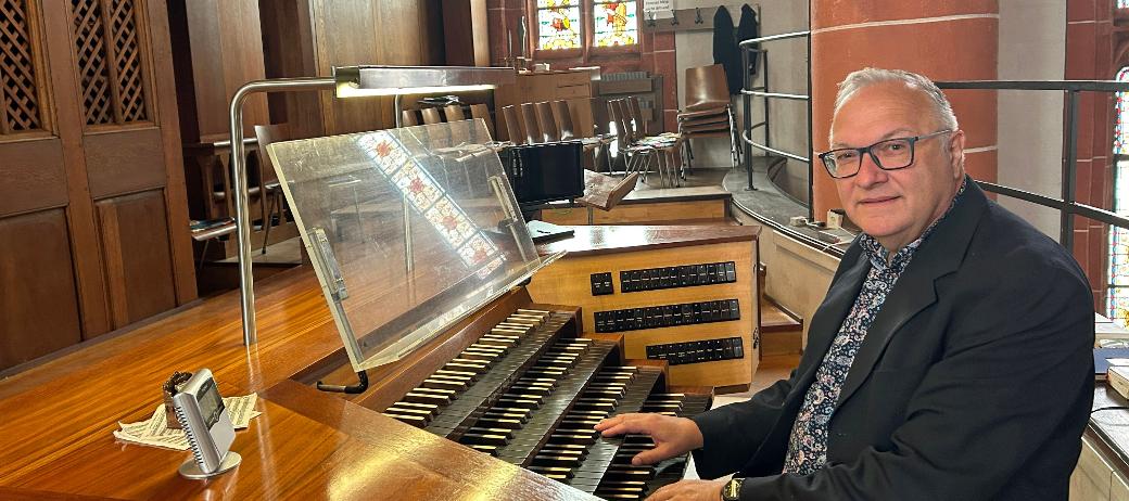 Ein Mann sitzt an der mehrmanualigen Orgel einer Kirche und hat die Hände auf den Tasten. Der hölzerne Spieltisch mit Notenpult und Registerschaltern steht auf einer Empore vor dem Orgelgehäuse mit sichtbaren Pfeifen. Im Hintergrund fallen Lichtstrahlen durch hohe Buntglasfenster, daneben sind Stuhlreihen und eine rötliche Steinsäule mit einer Heiligenfigur zu sehen. Die warme Holzoptik der Orgel prägt das Bild.