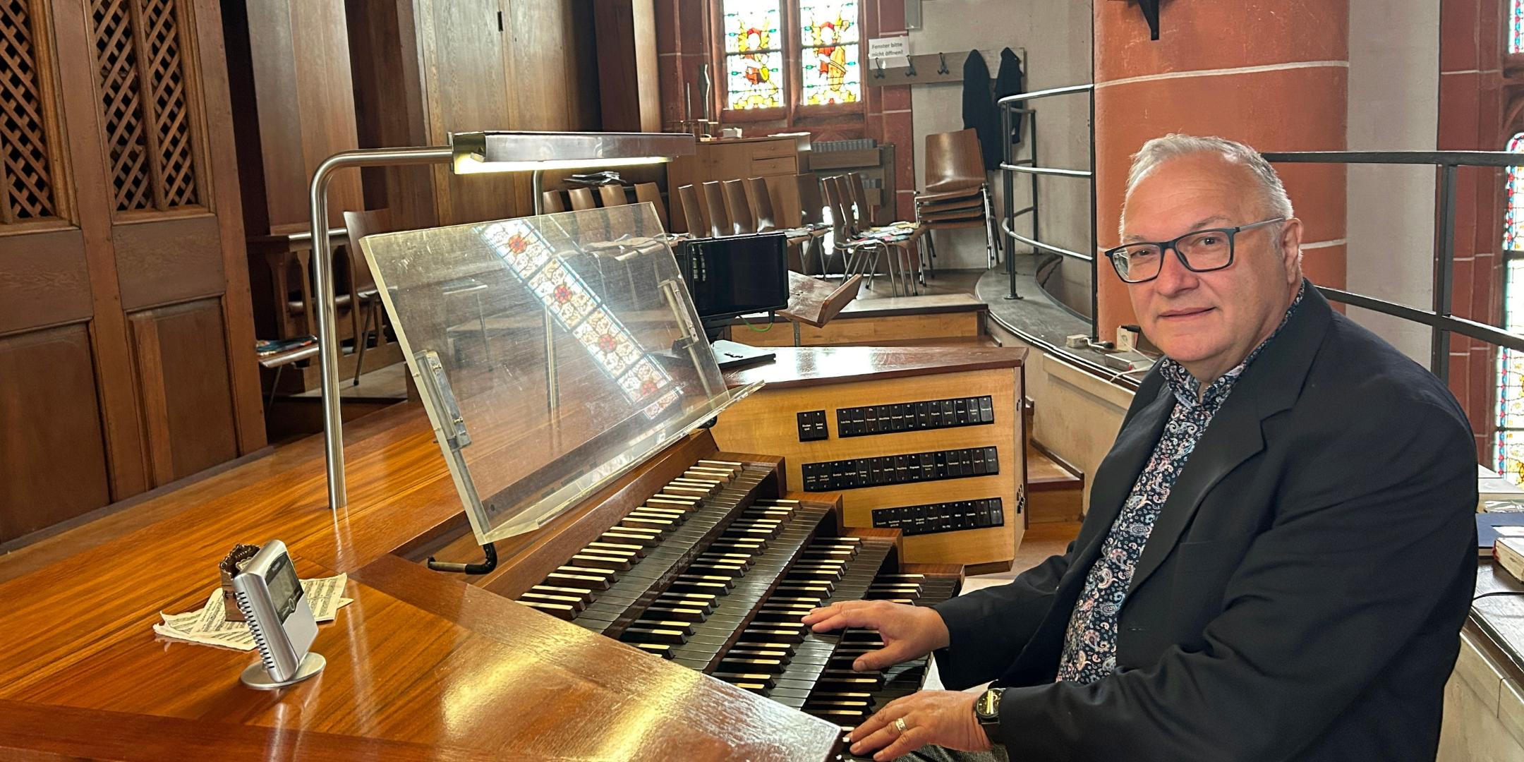 Ein Mann sitzt an der mehrmanualigen Orgel einer Kirche und hat die Hände auf den Tasten. Der hölzerne Spieltisch mit Notenpult und Registerschaltern steht auf einer Empore vor dem Orgelgehäuse mit sichtbaren Pfeifen. Im Hintergrund fallen Lichtstrahlen durch hohe Buntglasfenster, daneben sind Stuhlreihen und eine rötliche Steinsäule mit einer Heiligenfigur zu sehen. Die warme Holzoptik der Orgel prägt das Bild.