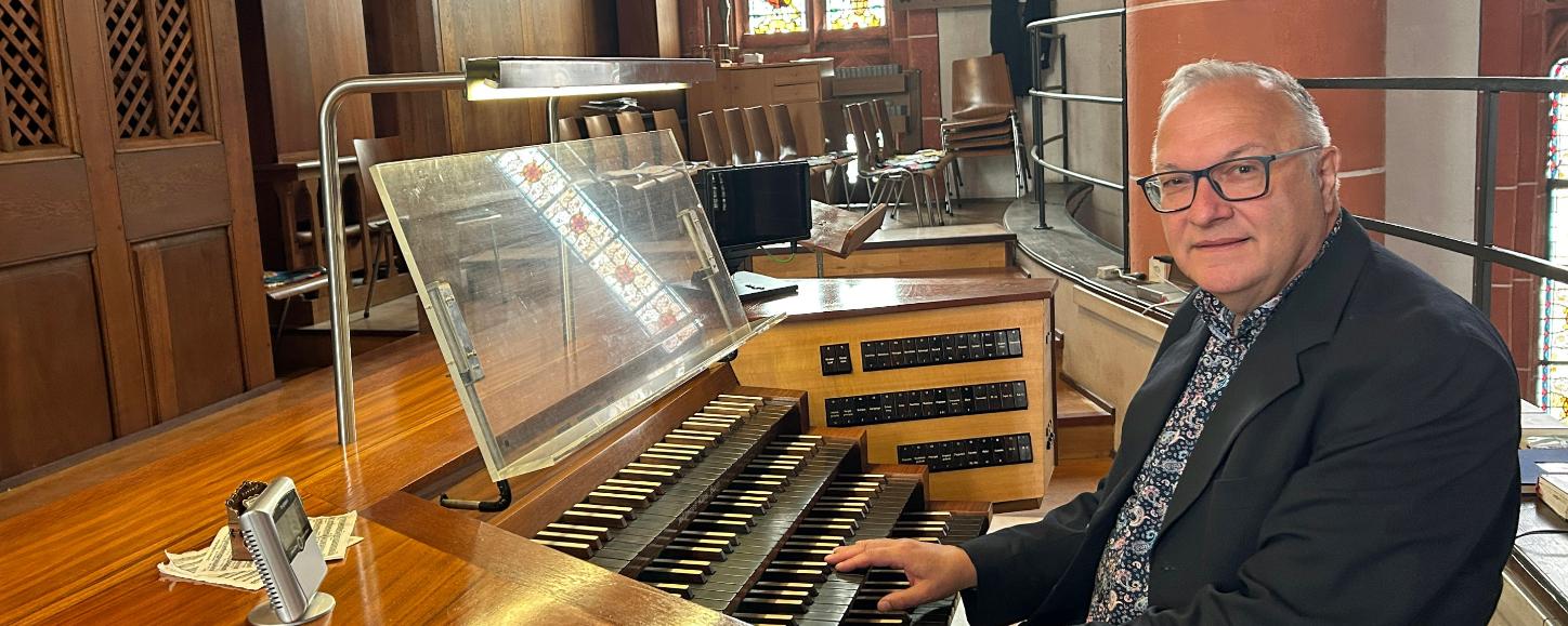 Ein Mann sitzt an der mehrmanualigen Orgel einer Kirche und hat die Hände auf den Tasten. Der hölzerne Spieltisch mit Notenpult und Registerschaltern steht auf einer Empore vor dem Orgelgehäuse mit sichtbaren Pfeifen. Im Hintergrund fallen Lichtstrahlen durch hohe Buntglasfenster, daneben sind Stuhlreihen und eine rötliche Steinsäule mit einer Heiligenfigur zu sehen. Die warme Holzoptik der Orgel prägt das Bild.