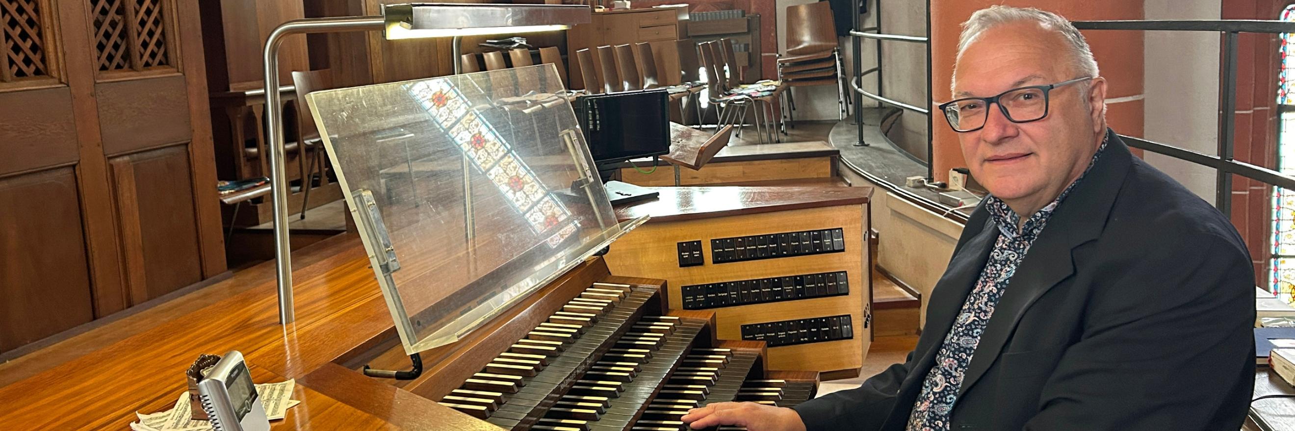 Ein Mann sitzt an der mehrmanualigen Orgel einer Kirche und hat die Hände auf den Tasten. Der hölzerne Spieltisch mit Notenpult und Registerschaltern steht auf einer Empore vor dem Orgelgehäuse mit sichtbaren Pfeifen. Im Hintergrund fallen Lichtstrahlen durch hohe Buntglasfenster, daneben sind Stuhlreihen und eine rötliche Steinsäule mit einer Heiligenfigur zu sehen. Die warme Holzoptik der Orgel prägt das Bild.
