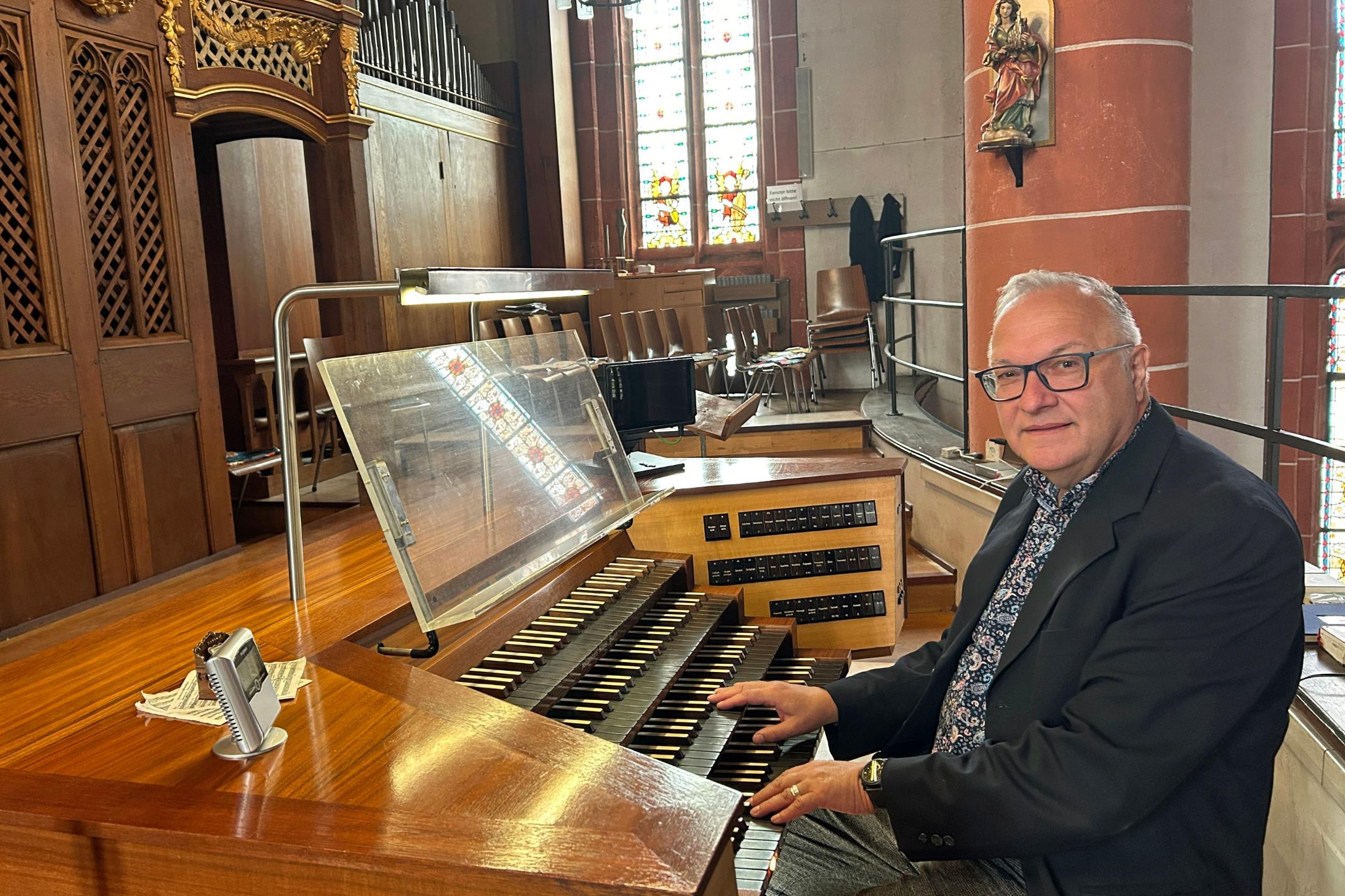 Ein Mann sitzt an der mehrmanualigen Orgel einer Kirche und hat die Hände auf den Tasten. Der hölzerne Spieltisch mit Notenpult und Registerschaltern steht auf einer Empore vor dem Orgelgehäuse mit sichtbaren Pfeifen. Im Hintergrund fallen Lichtstrahlen durch hohe Buntglasfenster, daneben sind Stuhlreihen und eine rötliche Steinsäule mit einer Heiligenfigur zu sehen. Die warme Holzoptik der Orgel prägt das Bild.