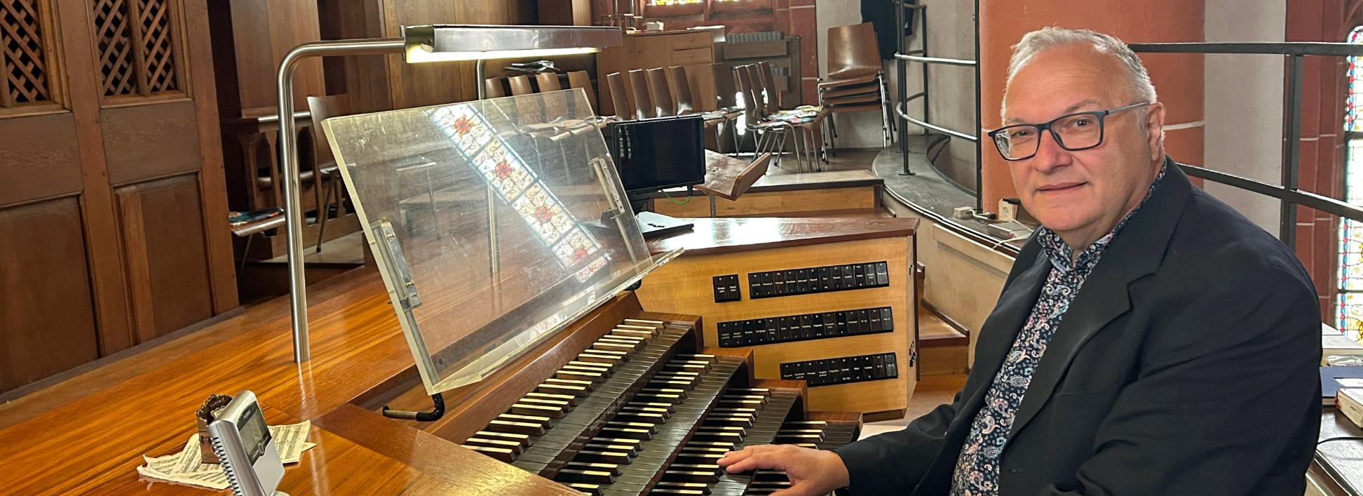 Ein Mann sitzt an der mehrmanualigen Orgel einer Kirche und hat die Hände auf den Tasten. Der hölzerne Spieltisch mit Notenpult und Registerschaltern steht auf einer Empore vor dem Orgelgehäuse mit sichtbaren Pfeifen. Im Hintergrund fallen Lichtstrahlen durch hohe Buntglasfenster, daneben sind Stuhlreihen und eine rötliche Steinsäule mit einer Heiligenfigur zu sehen. Die warme Holzoptik der Orgel prägt das Bild.