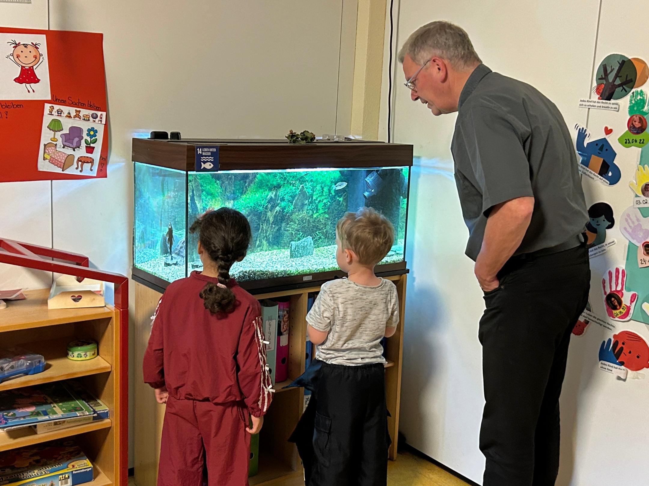Zwei Kinder zeigen Bischofsvikar Ulrich von Plettenberg das Aquarium und die Welse im Gruppenraum.