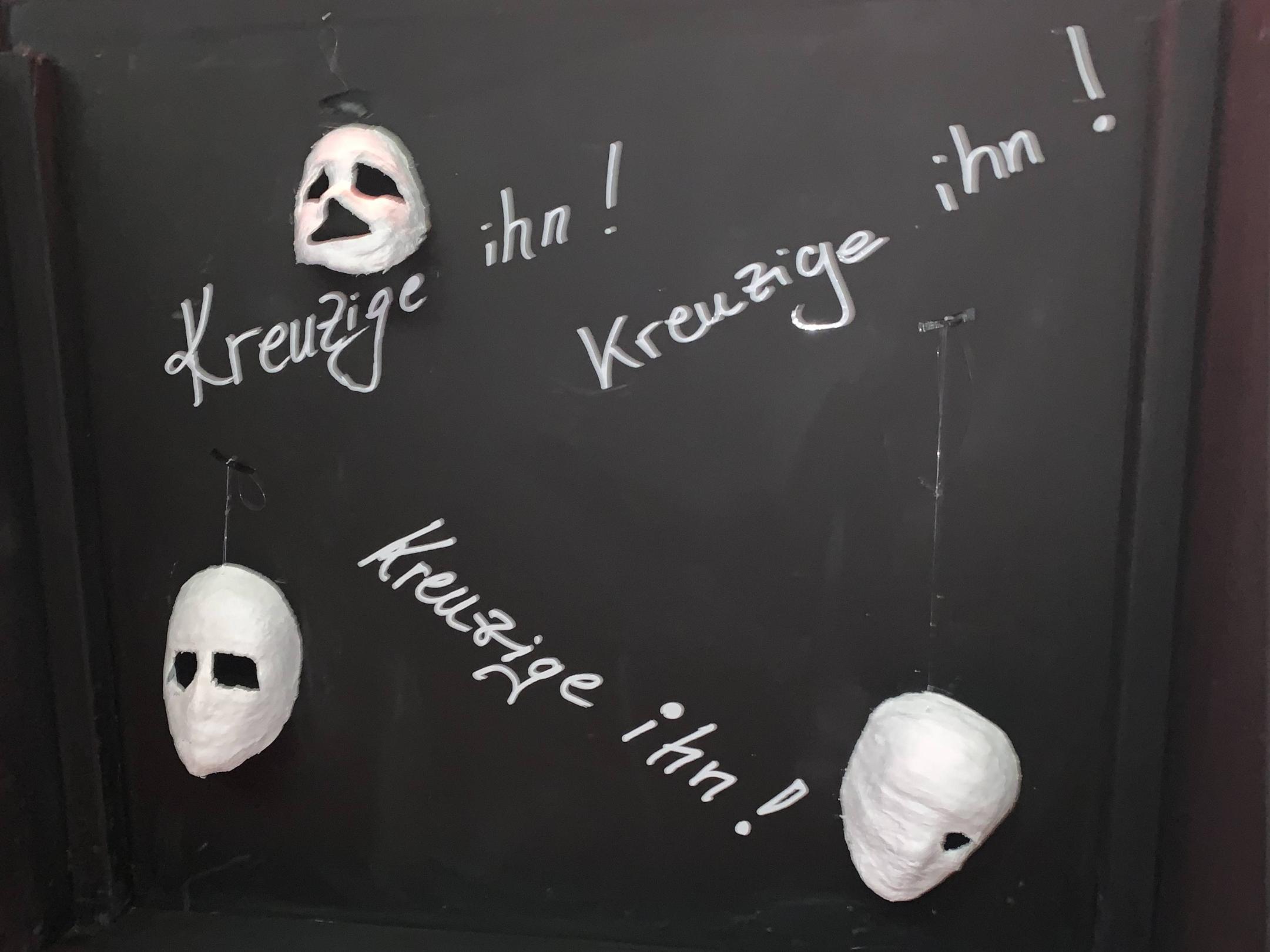 Schwarze Fläche mit weißer 'Kreizige ihn!'-Aufschrift und weißen Masken
