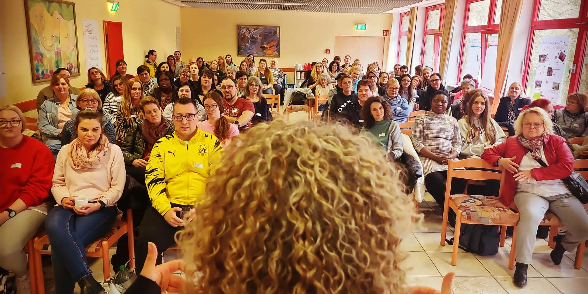 viele Menschen in einem Raum. Im Vordergrund Frau mit Lockenkopf.