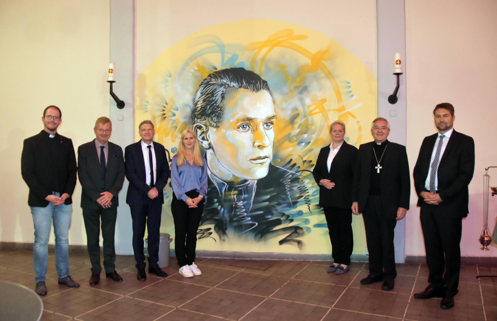 Jugendpfarrer Thomas Hufschmidt, der französische Generalkonsul Sebastién Girard, Stephan Toscani (Mitglied im Beirat der Weiße Rose Stiftung e.V.), , Juliane Baez (Großnichte Willi Grafs), Landtagspräsidentin Heike Becker, Weihbischof Franz Josef Gebert und Oberbürgermeister Uwe Conradt (von links) vor dem Porträt Willi Grafs des Streetart-Künstlers Christian Guémy in der Jugendkirche.
