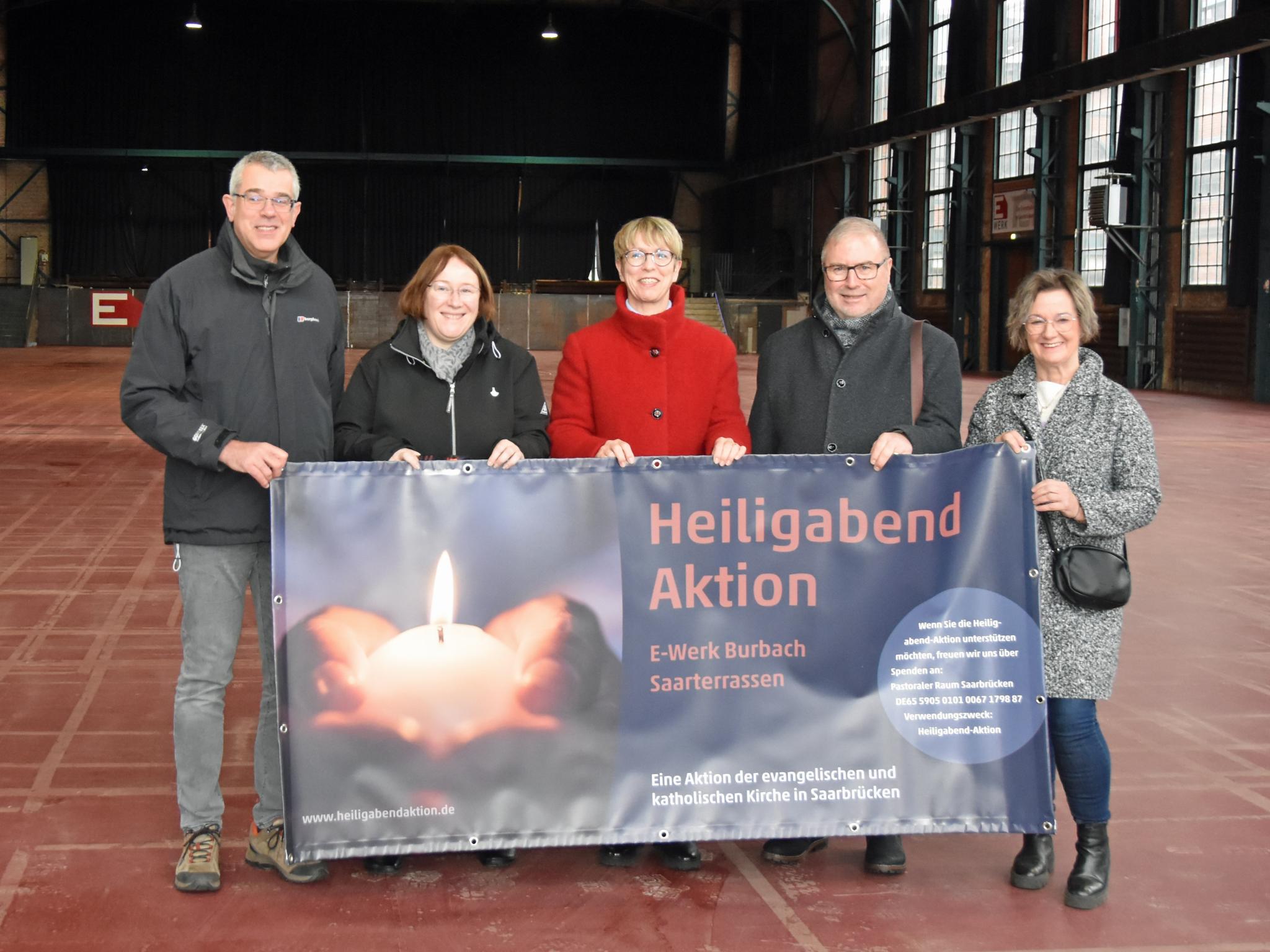 Fünf Personen stehen in einer Halle und halten das Plakat zur Heiligabend-Aktion.