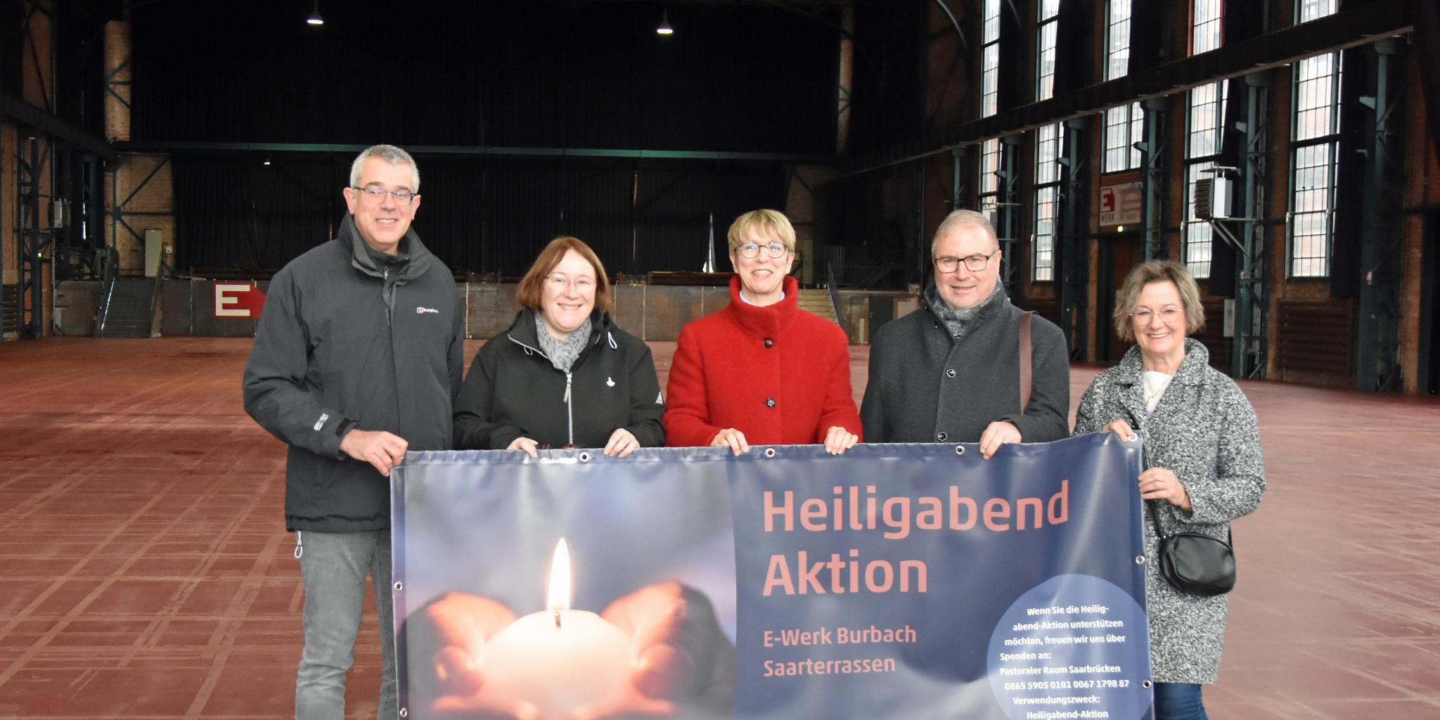 Fünf Personen stehen in einer Halle und halten das Plakat zur Heiligabend-Aktion.