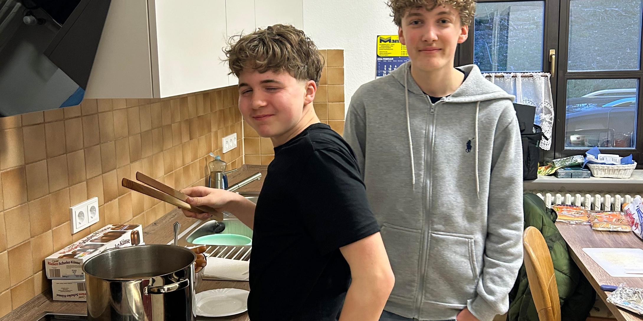 Zwei Jugendliche stehen in einer Küche. Eine Person steht am Herd und rührt mit Küchenzangen in einem großen Edelstahlkochtopf auf der Arbeitsplatte. Daneben liegen ein Topfdeckel und Zutaten wie Hotdog-Brötchen. Die zweite Person steht leicht seitlich dahinter, mit den Händen in den Hosentaschen. Im Hintergrund sind Küchenschränke, ein Fenster, ein Esstisch mit Stühlen und weitere Lebensmittel zu sehen.