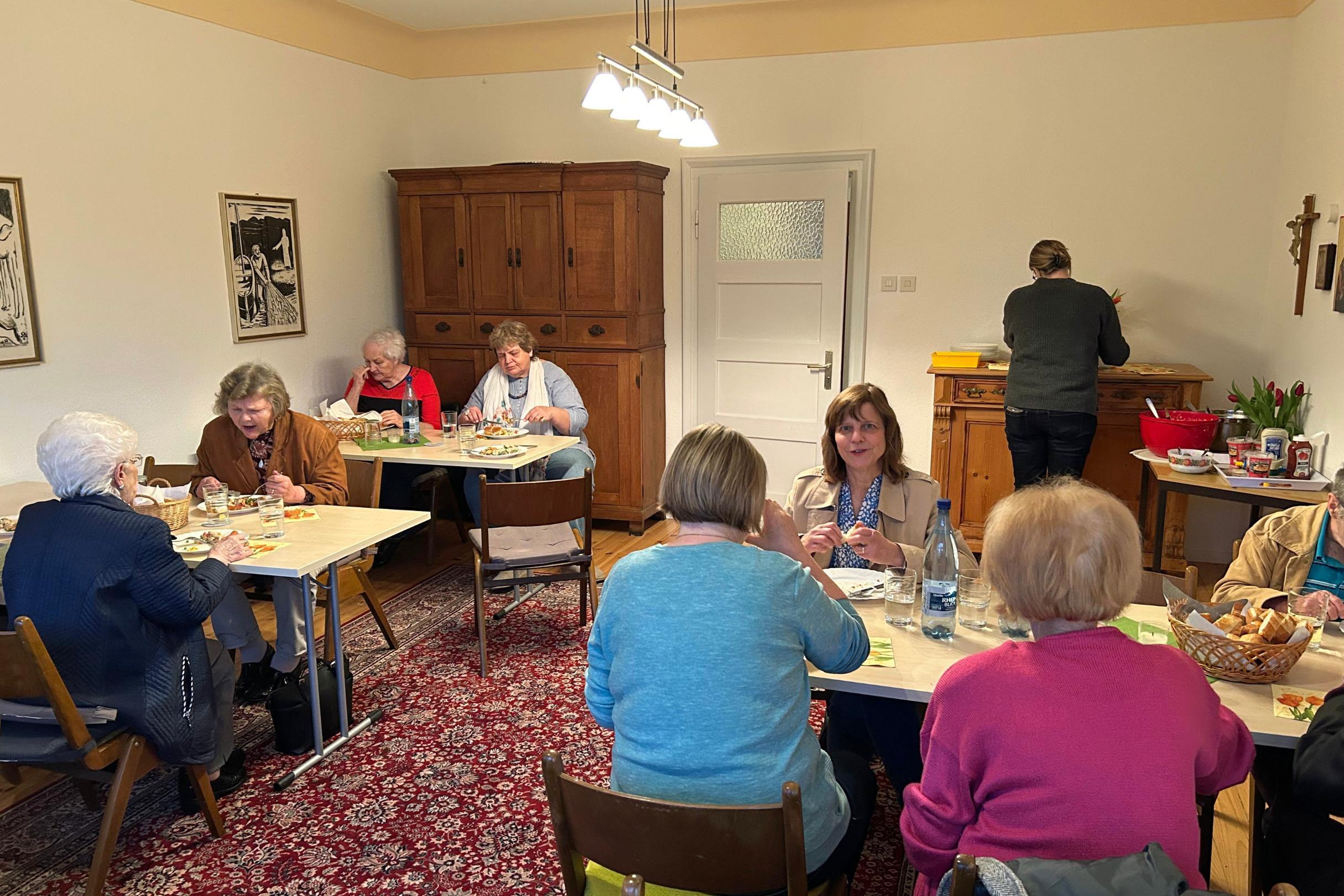 In einem gemütlichen Gemeinderaum sitzen mehrere Personen an kleinen Tischen und essen gemeinsam. Die Tische sind mit einfachen Tischdecken gedeckt und mit Getränken, Tellern und Besteck versehen. Auf dem roten Teppichboden stehen verschiedene Holzstühle. An der rechten Seite des Raums befindet sich ein Sideboard mit Lebensmitteln und Geschirr, an dem eine Person arbeitet. Im Hintergrund stehen ein großer Holzschrank und ein weißer Türbereich. Die Wände sind hell und mit zwei gerahmten Bildern dekoriert.