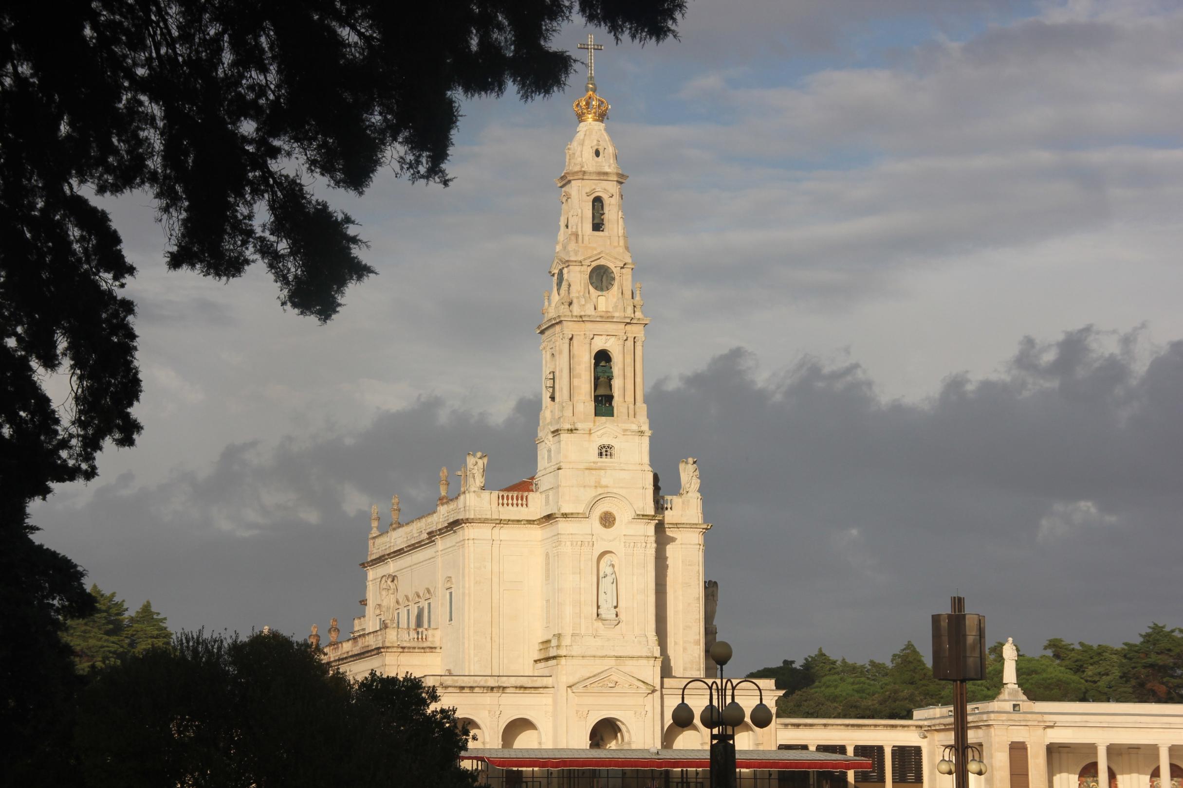 Sonnenbeschienene Basilika unserer lieben Frau des Rosenkranzes in Fátima unter wolkigem Himmel, Bäume im Vordergrund.