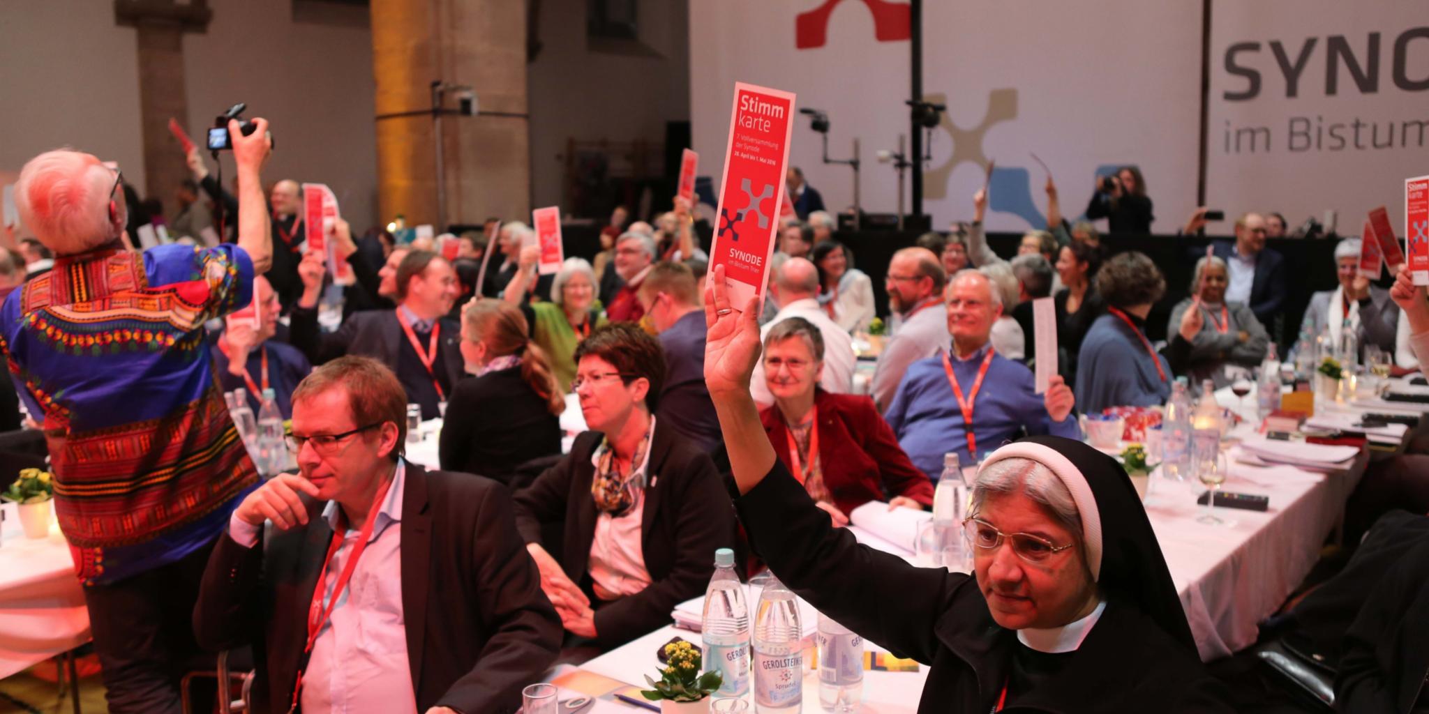Auf dem Bild sind zahlreiche Menschen in einem großen Saal bei einer Veranstaltung zu sehen, viele von ihnen halten rote Karten mit der Aufschrift „Stimmkarte“ in die Höhe. Im Vordergrund sitzt eine Ordensschwester, die ebenfalls eine Stimmkarte hochhält, während andere Teilnehmer aufmerksam blicken oder diskutieren. Im Hintergrund ist ein großes Transparent mit der Aufschrift „Synode im Bistum“ sichtbar. (Alt-Text erstellt mit KI-Unterstützung)