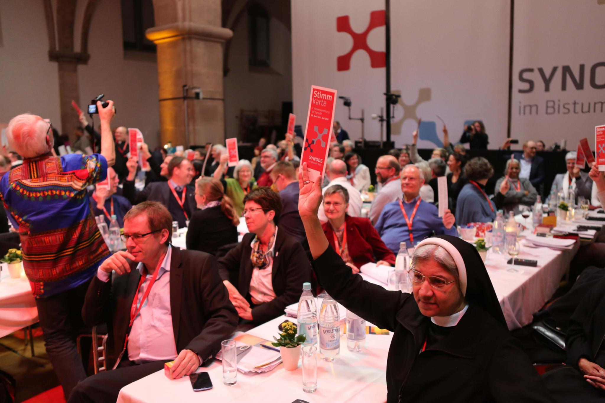 Auf dem Bild sind zahlreiche Menschen in einem großen Saal bei einer Veranstaltung zu sehen, viele von ihnen halten rote Karten mit der Aufschrift „Stimmkarte“ in die Höhe. Im Vordergrund sitzt eine Ordensschwester, die ebenfalls eine Stimmkarte hochhält, während andere Teilnehmer aufmerksam blicken oder diskutieren. Im Hintergrund ist ein großes Transparent mit der Aufschrift „Synode im Bistum“ sichtbar. (Alt-Text erstellt mit KI-Unterstützung)