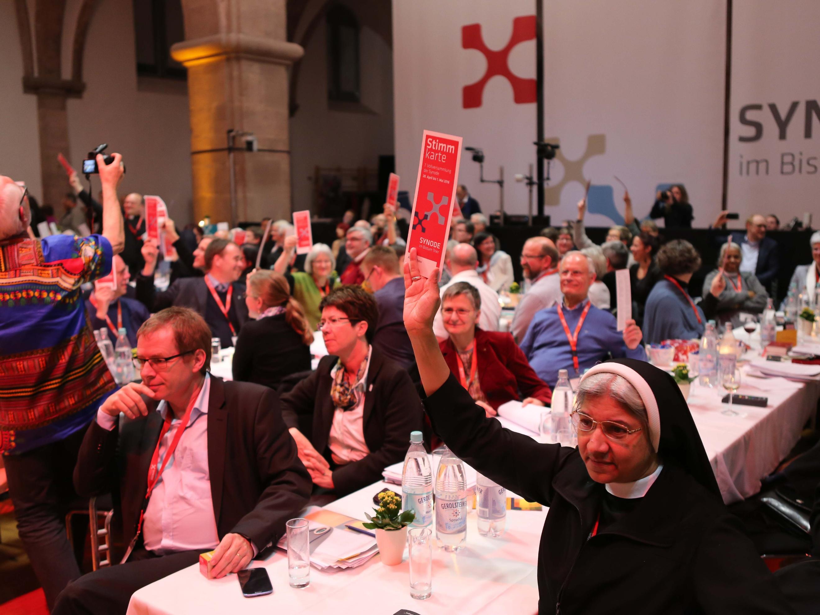 Auf dem Bild sind zahlreiche Menschen in einem großen Saal bei einer Veranstaltung zu sehen, viele von ihnen halten rote Karten mit der Aufschrift „Stimmkarte“ in die Höhe. Im Vordergrund sitzt eine Ordensschwester, die ebenfalls eine Stimmkarte hochhält, während andere Teilnehmer aufmerksam blicken oder diskutieren. Im Hintergrund ist ein großes Transparent mit der Aufschrift „Synode im Bistum“ sichtbar. (Alt-Text erstellt mit KI-Unterstützung)