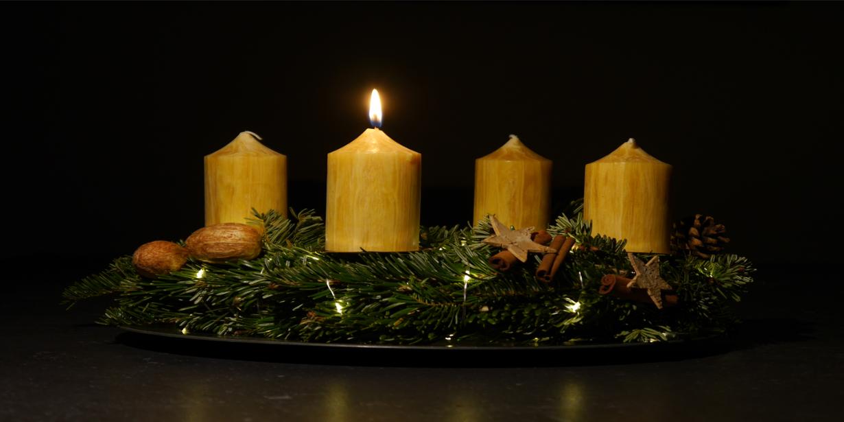 Das Foto zeigt in Großaufnahme einen Adventskranz vor dunklem Hintergrund. Der Adventskranz hat honigfarbene Kerzen, von denen eine brennt.
