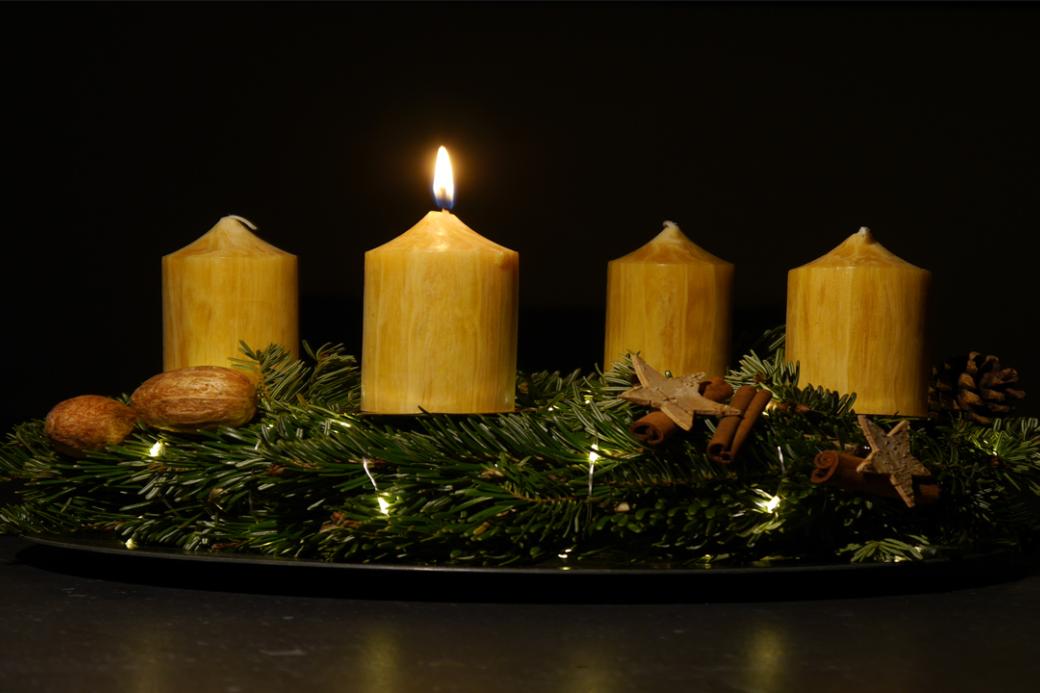 Das Foto zeigt in Großaufnahme einen Adventskranz vor dunklem Hintergrund. Der Adventskranz hat honigfarbene Kerzen, von denen eine brennt.