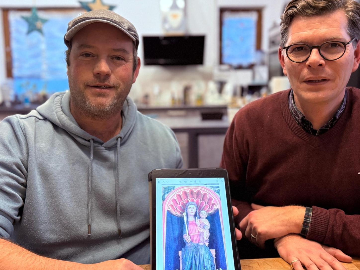 Zwei Männer an einem Tisch zeigen ein Tablet mit dem Foto einer Madonnen-Figur.