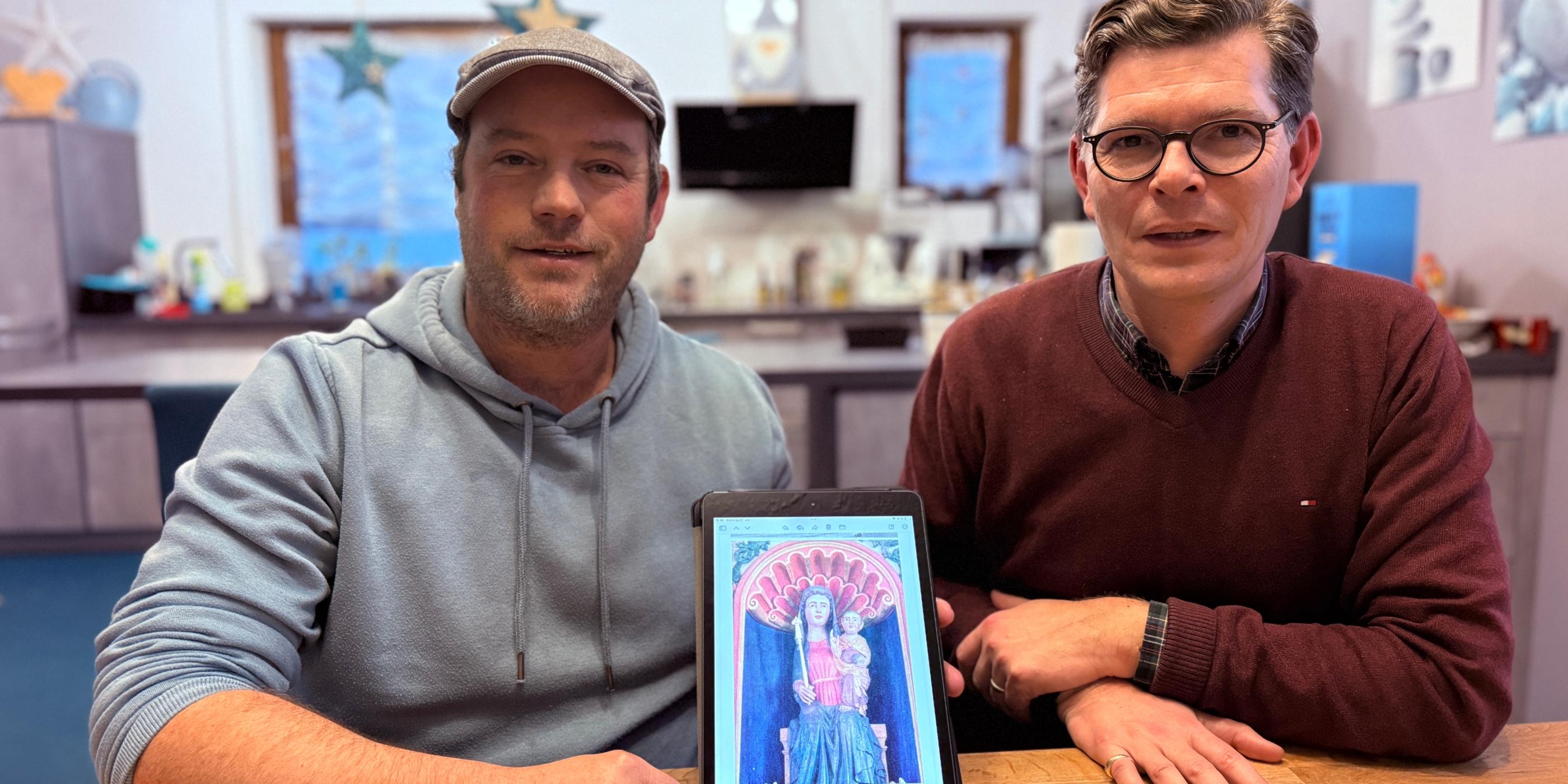 Zwei Männer an einem Tisch zeigen ein Tablet mit dem Foto einer Madonnen-Figur.