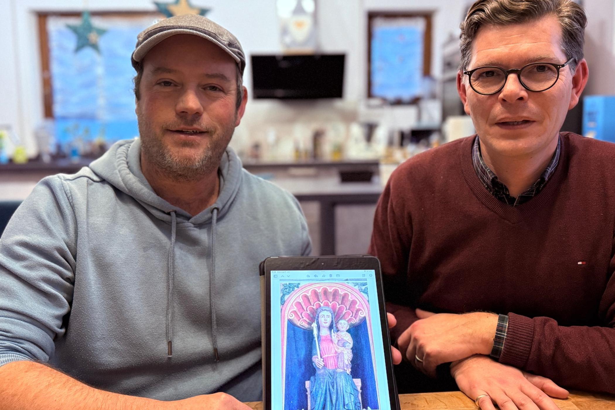 Zwei Männer an einem Tisch zeigen ein Tablet mit dem Foto einer Madonnen-Figur.