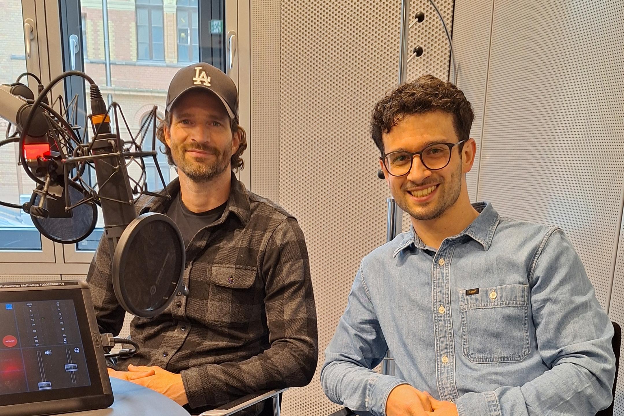 Zwei Männer sitzen lächelnd in einem modernen Radiostudio vor Mikrofonen. (KI-Text)