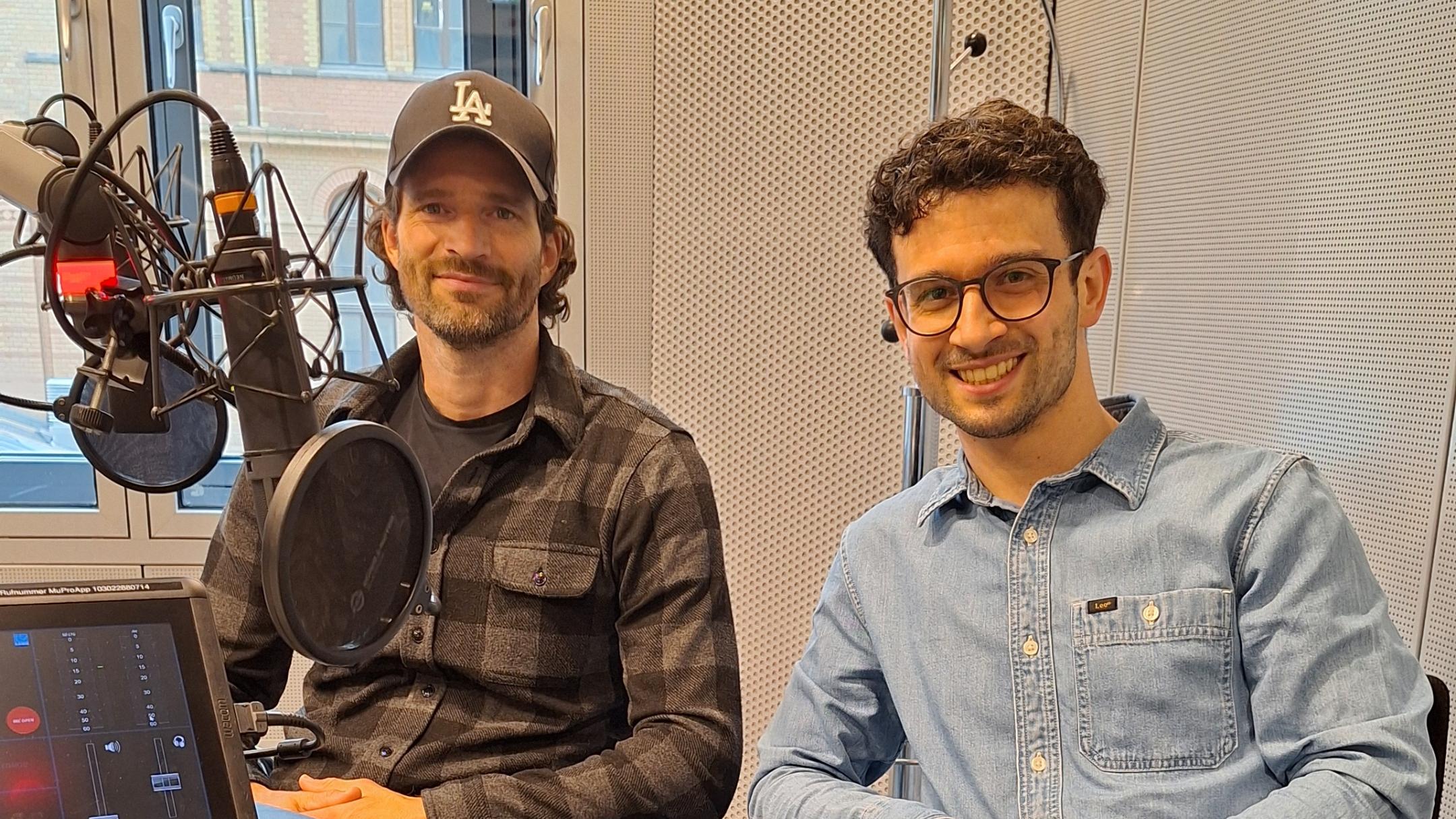 Zwei Männer sitzen lächelnd in einem modernen Radiostudio vor Mikrofonen. (KI-Text)