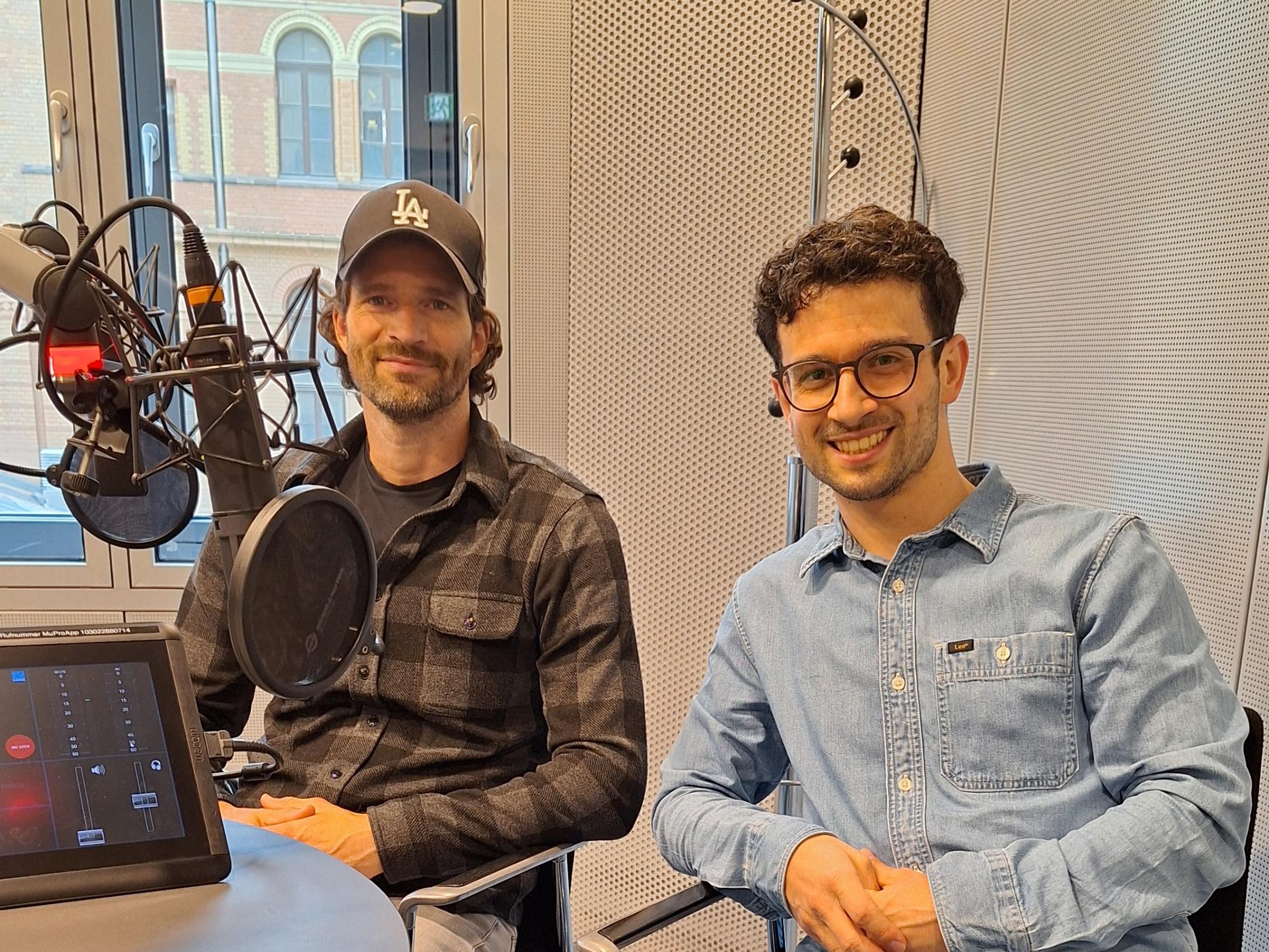 Zwei Männer sitzen lächelnd in einem modernen Radiostudio vor Mikrofonen. (KI-Text)