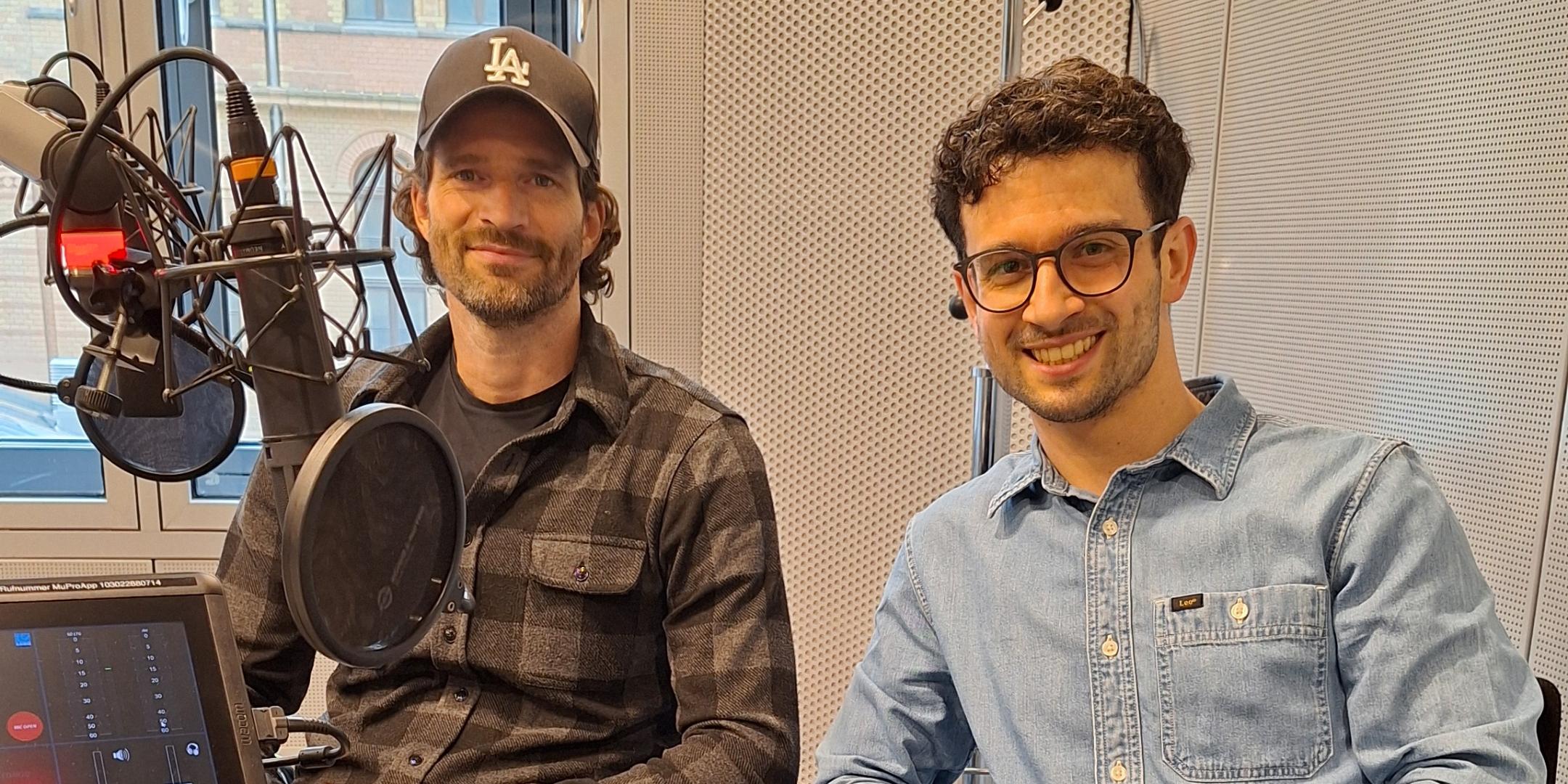Zwei Männer sitzen lächelnd in einem modernen Radiostudio vor Mikrofonen. (KI-Text)