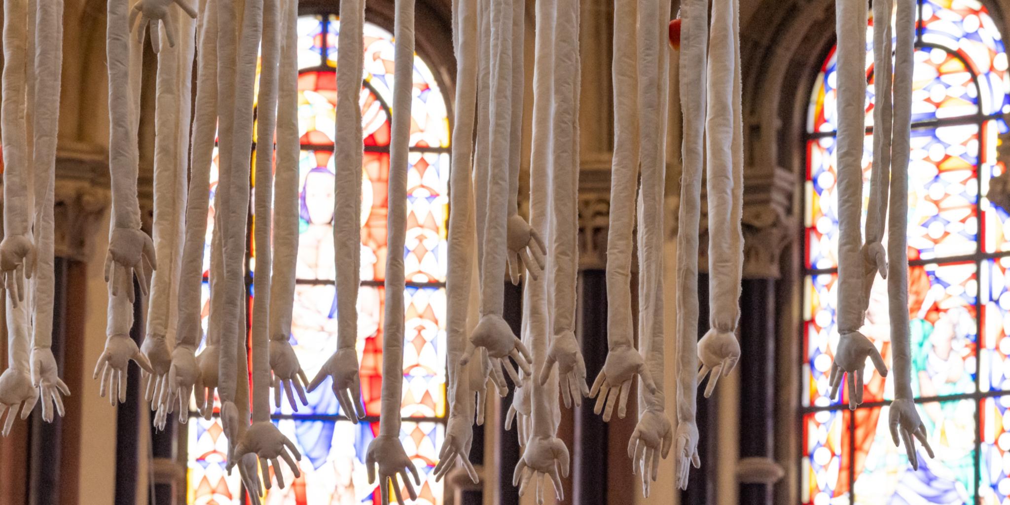 Herabhängende Hände und Arme aus weißem Leinen vor Kirchenfenstern.