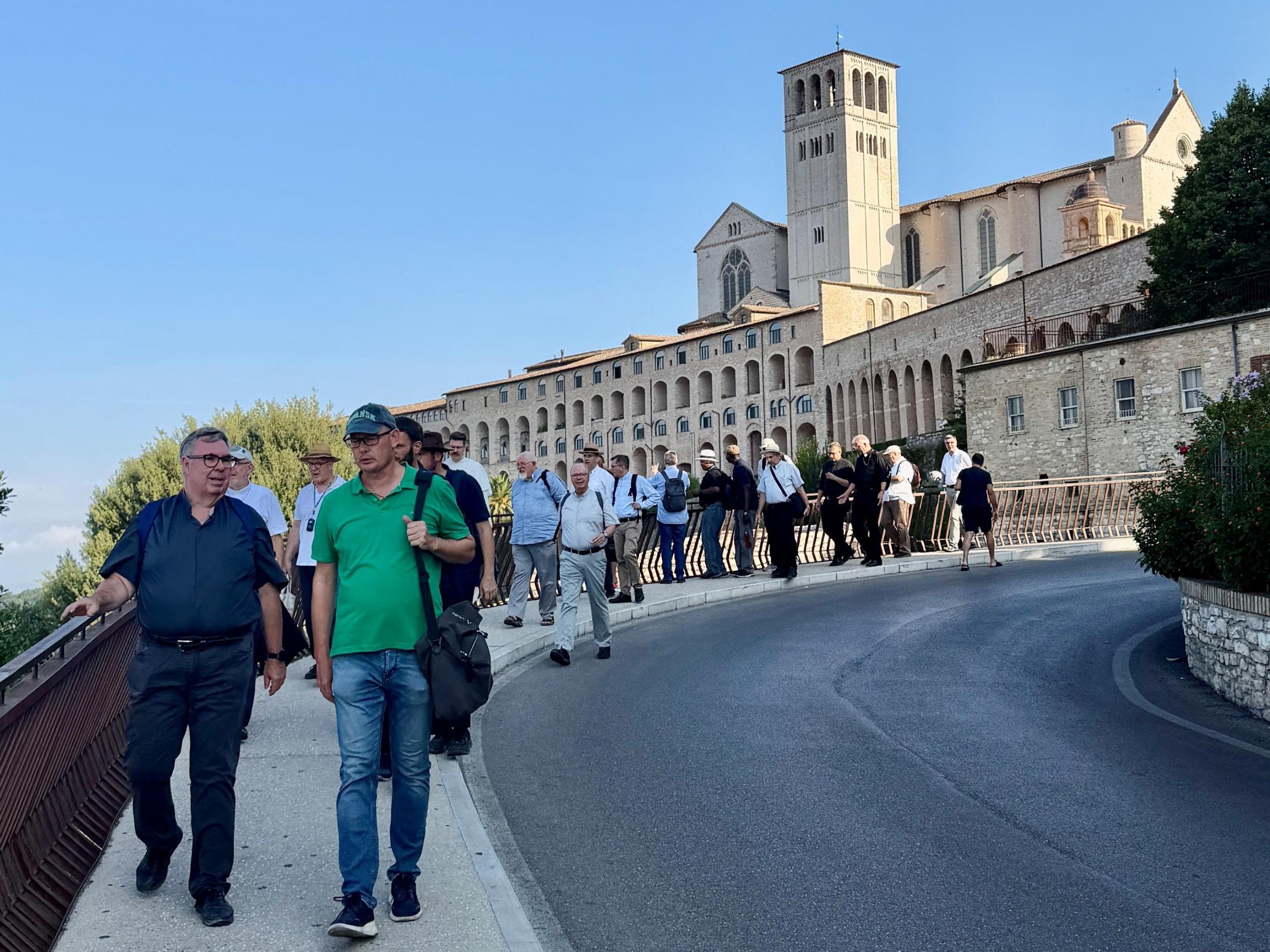 Priester spazieren hintereinander auf einem Gehweg, dahinter die Kirche von Assisi