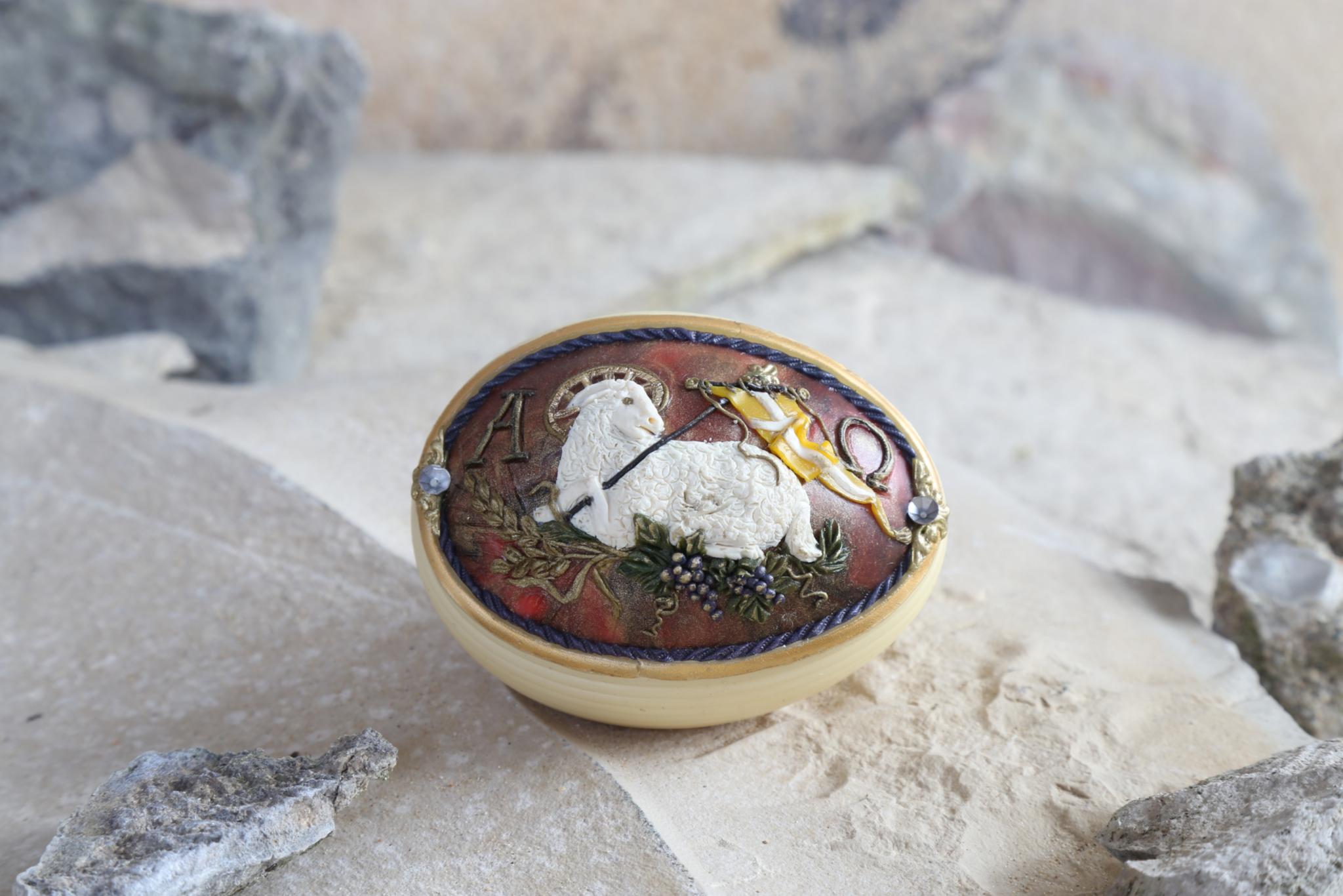 Ein dekoratives, ovales Ei liegt auf hellem, sandigem Untergrund zwischen einigen Steinen. Auf dem Ei befindet sich ein plastisch gestaltetes Relief eines weißen Lammes, das auf einem braun-rötlichen Hintergrund ruht. Neben dem Lamm ist eine goldene Standarte angebracht. Das Ei ist von einem dunklen Zierband umrahmt und wirkt handgefertigt und filigran.
