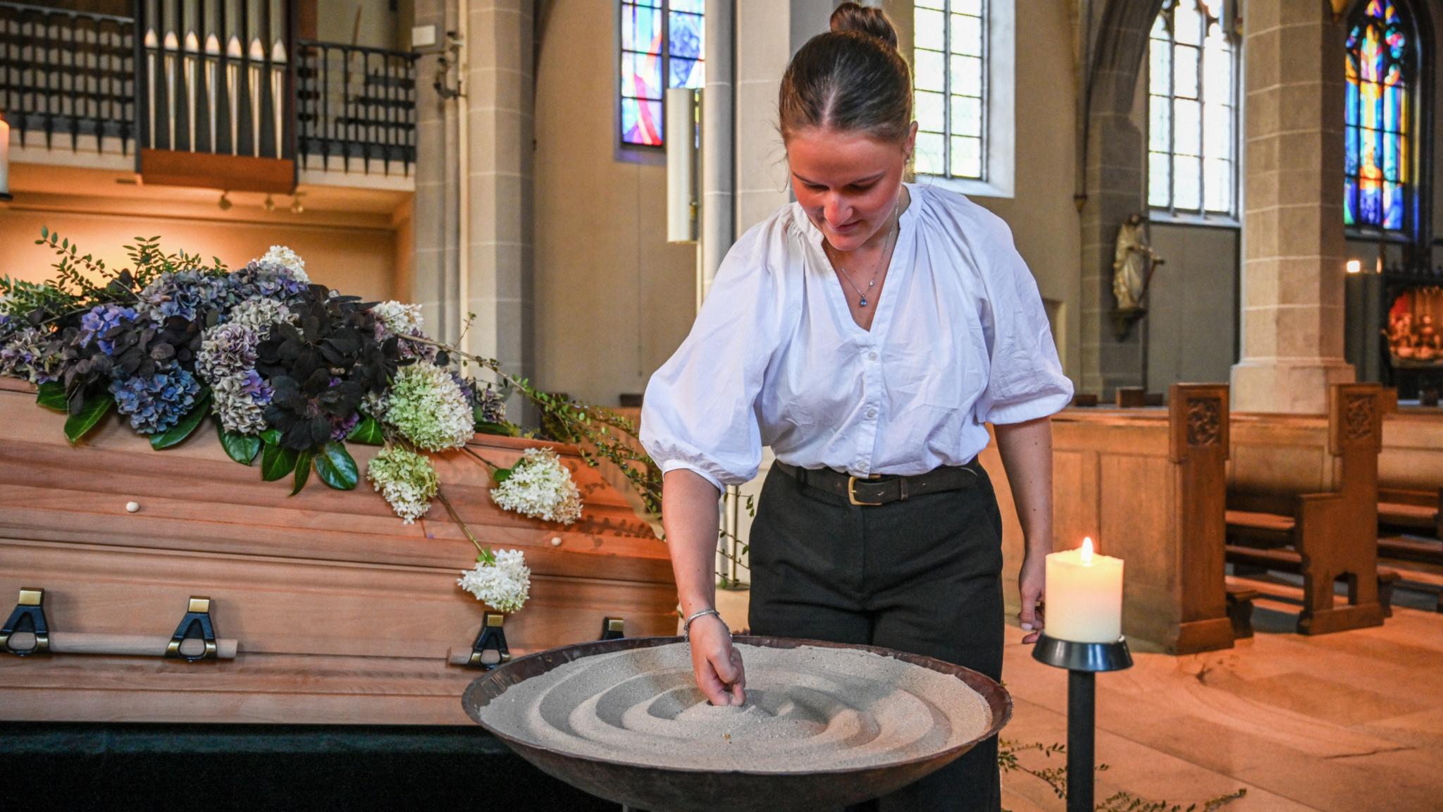 Eine Bestattering formt Sand in einer Schale, die vor einem geschmückten Sarg steht.